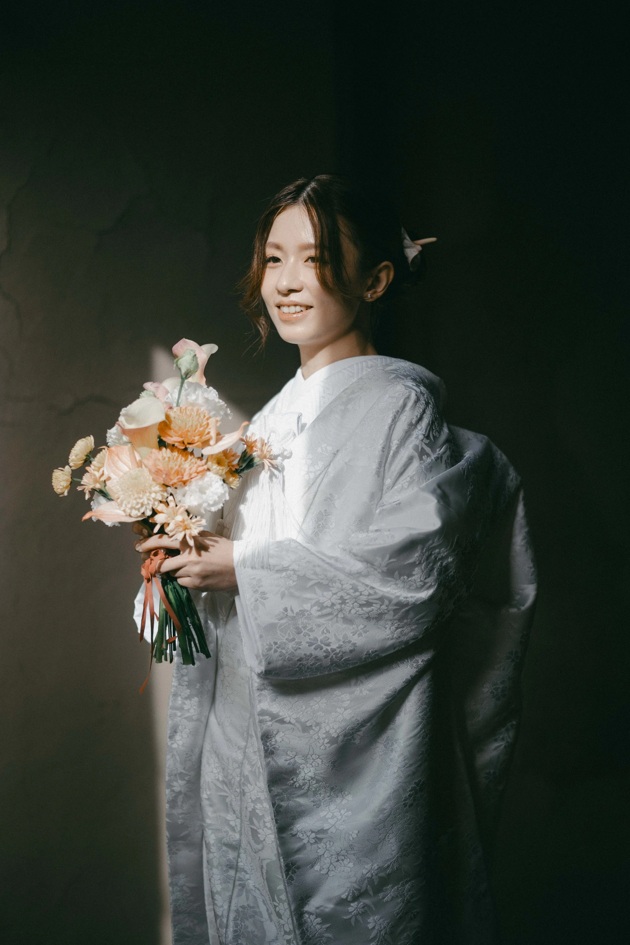 A woman in a traditional Japanese kimono holding a bouquet of flowers, standing in a dimly lit room with a dark background.
