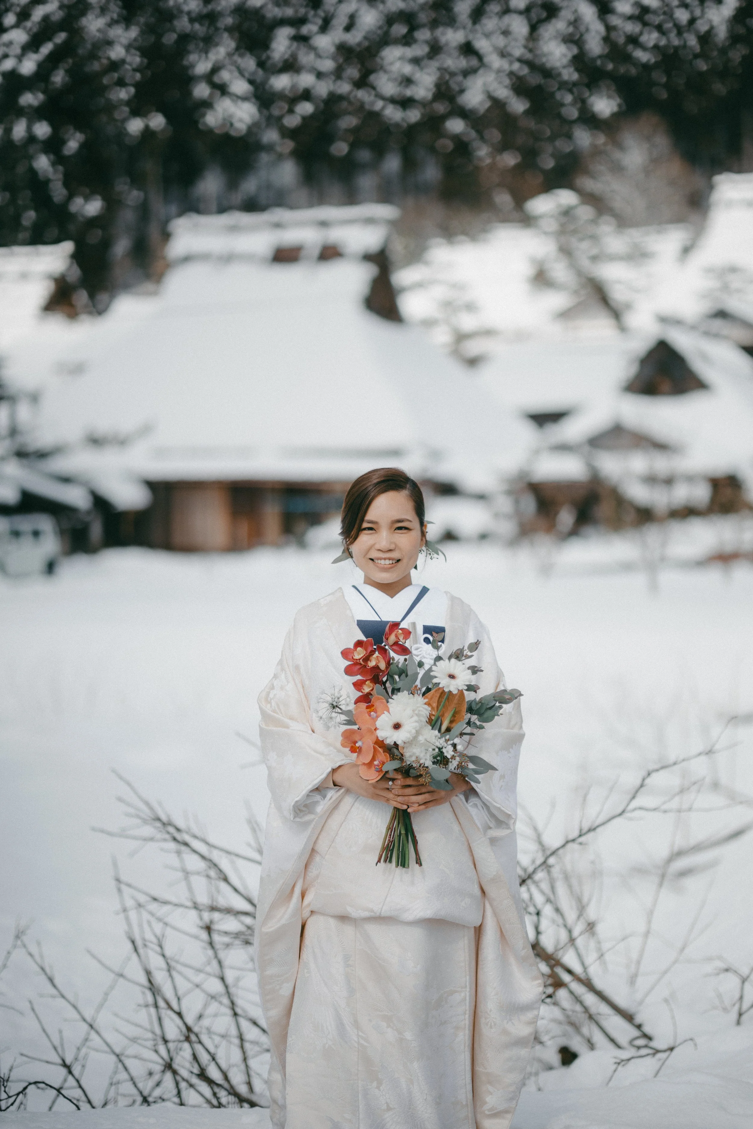 A woman in traditional Japanese wedding attire holding a bouquet of flowers, standing outdoors in a snowy landscape with traditional wooden buildings in the background.
