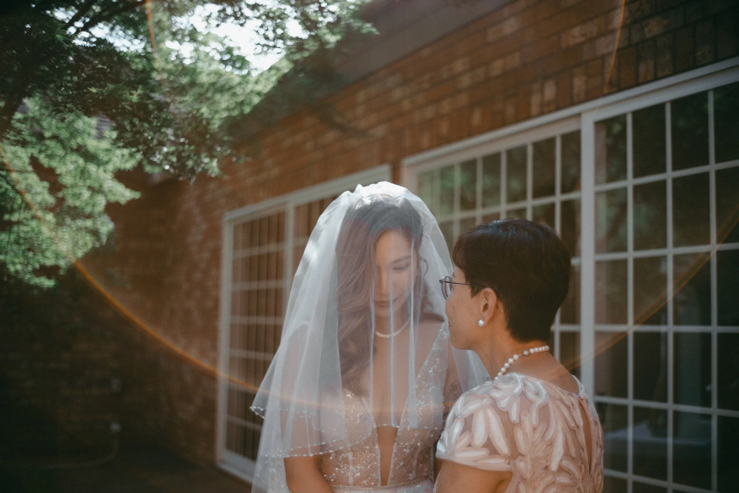 A bride in a wedding dress with a veil and a woman in a floral dress with pearl jewelry standing outside near a brick building with large windows during daytime.