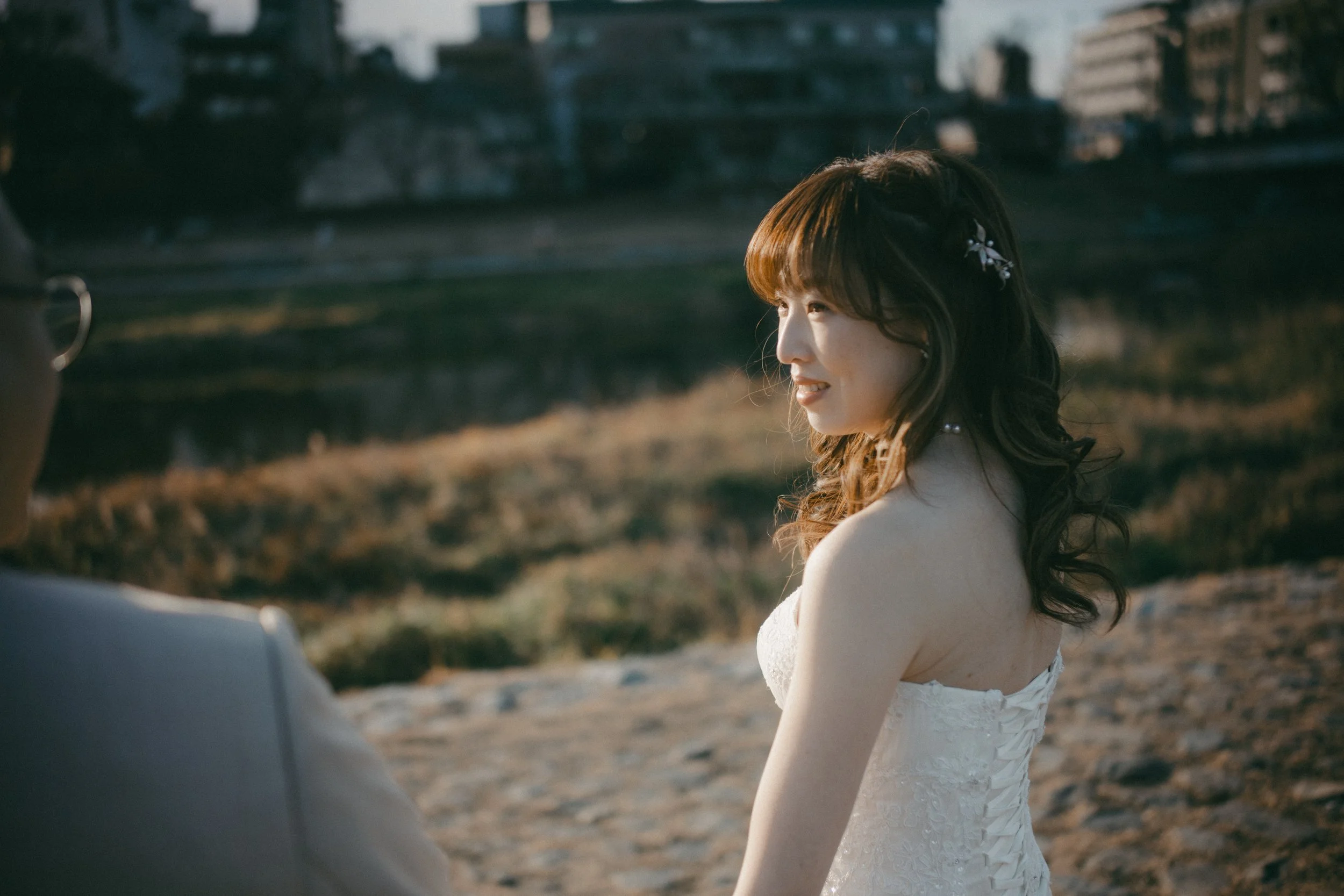 A bride and groom standing outdoors at sunset, with the bride wearing a white wedding dress and the groom in a suit, in a romantic moment by a stream.