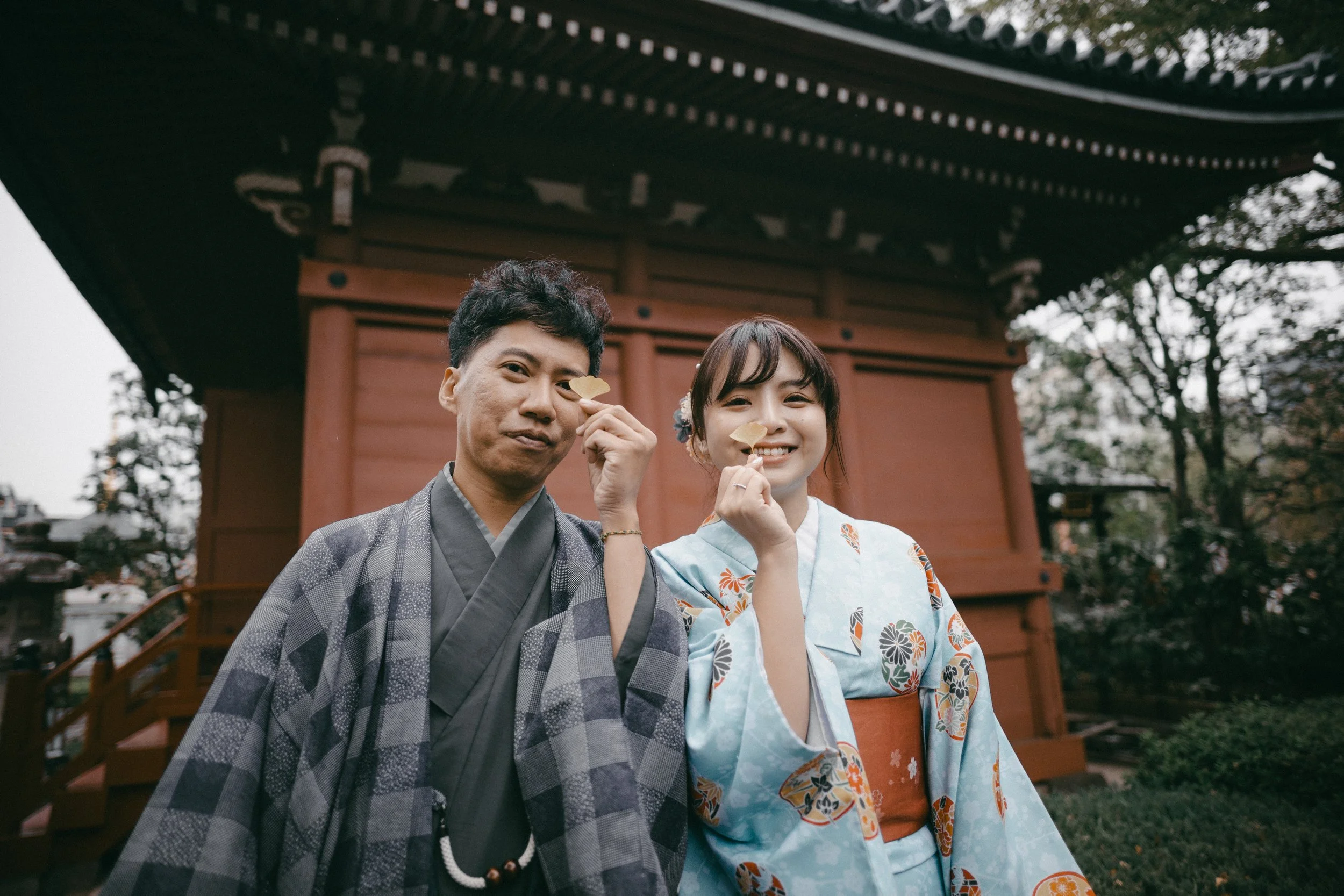 Two people in traditional Japanese clothing holding fan-shaped objects near a red temple in Japan.