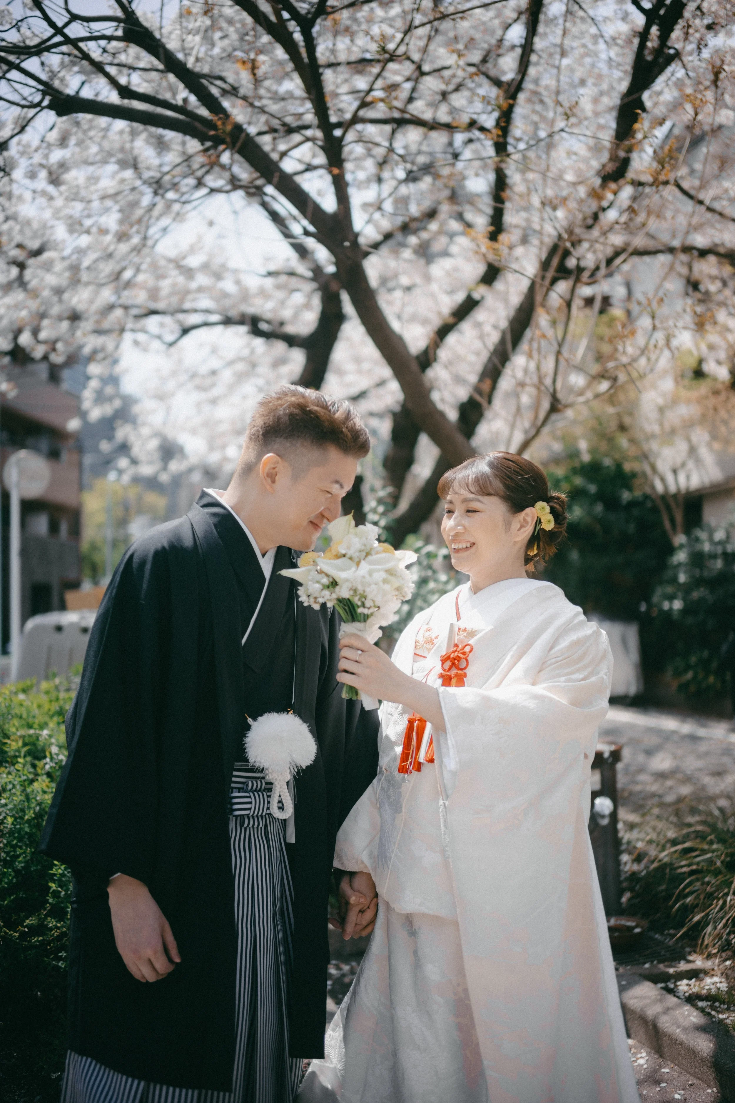 Couple in traditional Japanese wedding attire holding hands and smiling outdoors under cherry blossom trees.