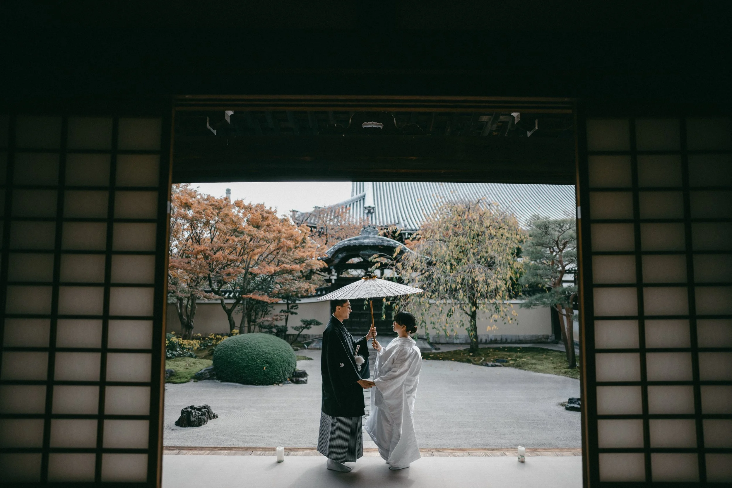 A couple dressed in traditional Japanese wedding attire sharing a moment in a Japanese garden, with one holding an umbrella over the other.
