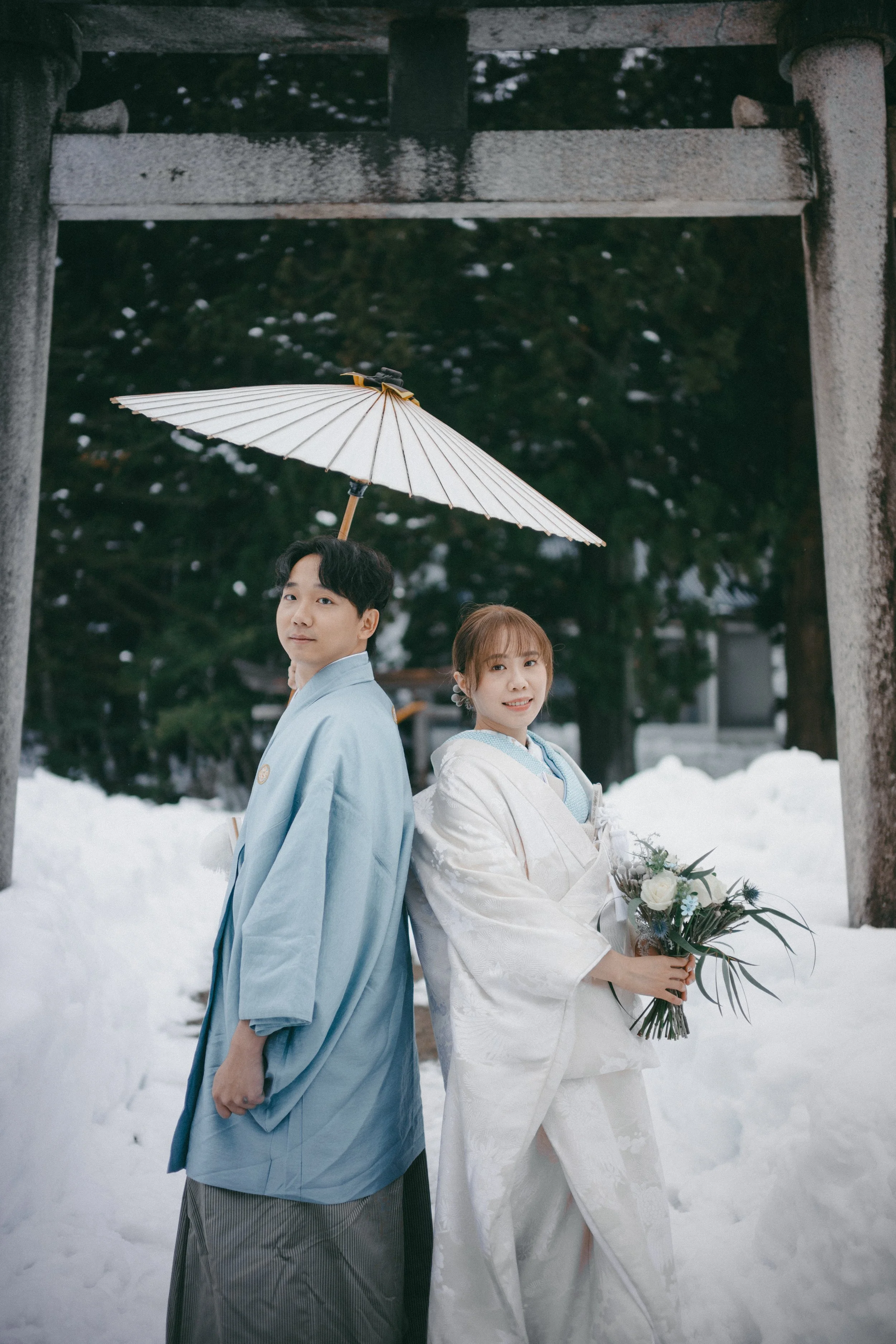 A couple dressed in traditional Japanese wedding attire standing back-to-back under a white umbrella, with snow on the ground and a torii gate in the background.