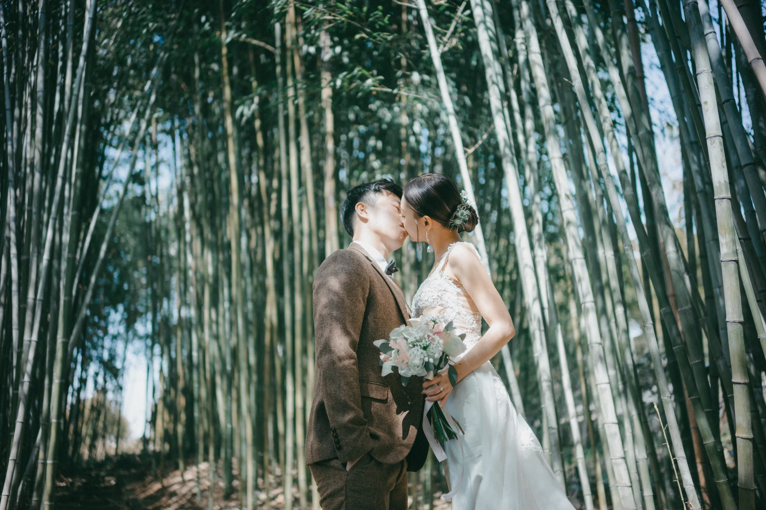 A bride and groom kiss in a bamboo forest, with the bride holding a bouquet of flowers.