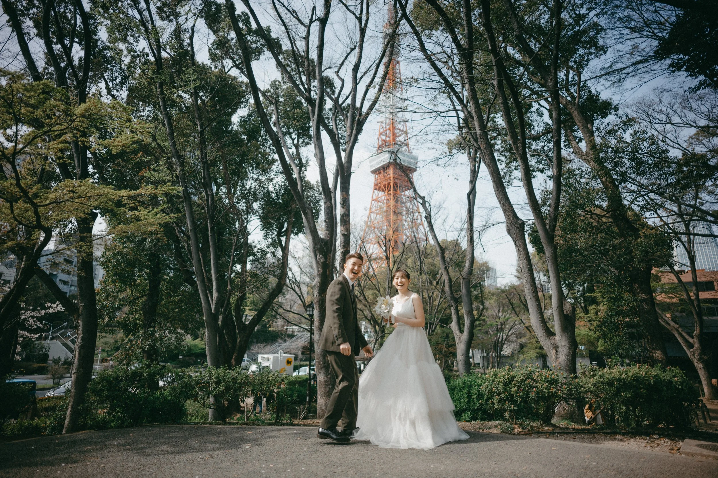A happy bride and groom in wedding attire standing outdoors surrounded by trees, with Tokyo Tower visible in the background.