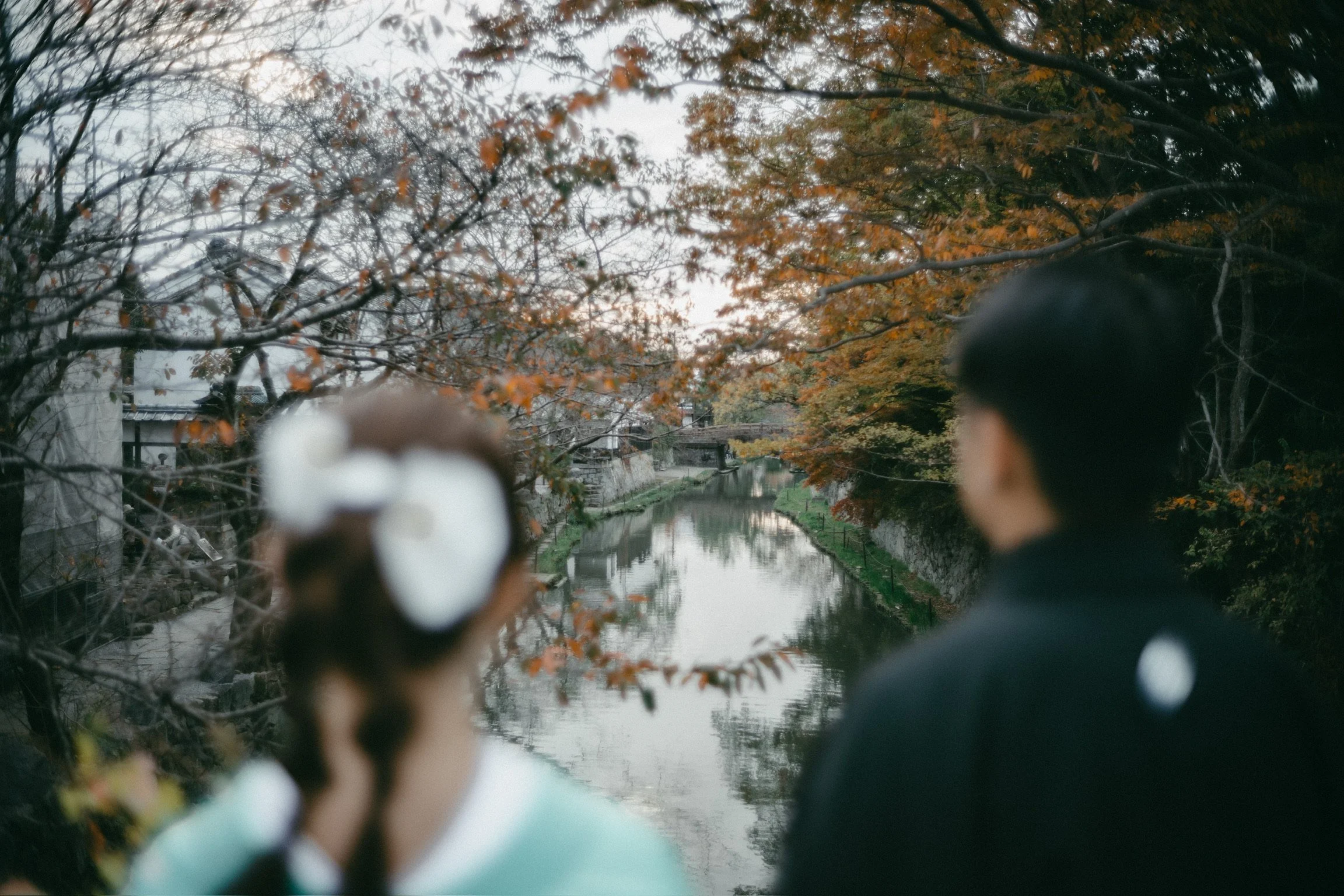 A couple is looking at a calm river surrounded by autumn trees with orange and brown leaves, possibly during late afternoon or early evening.