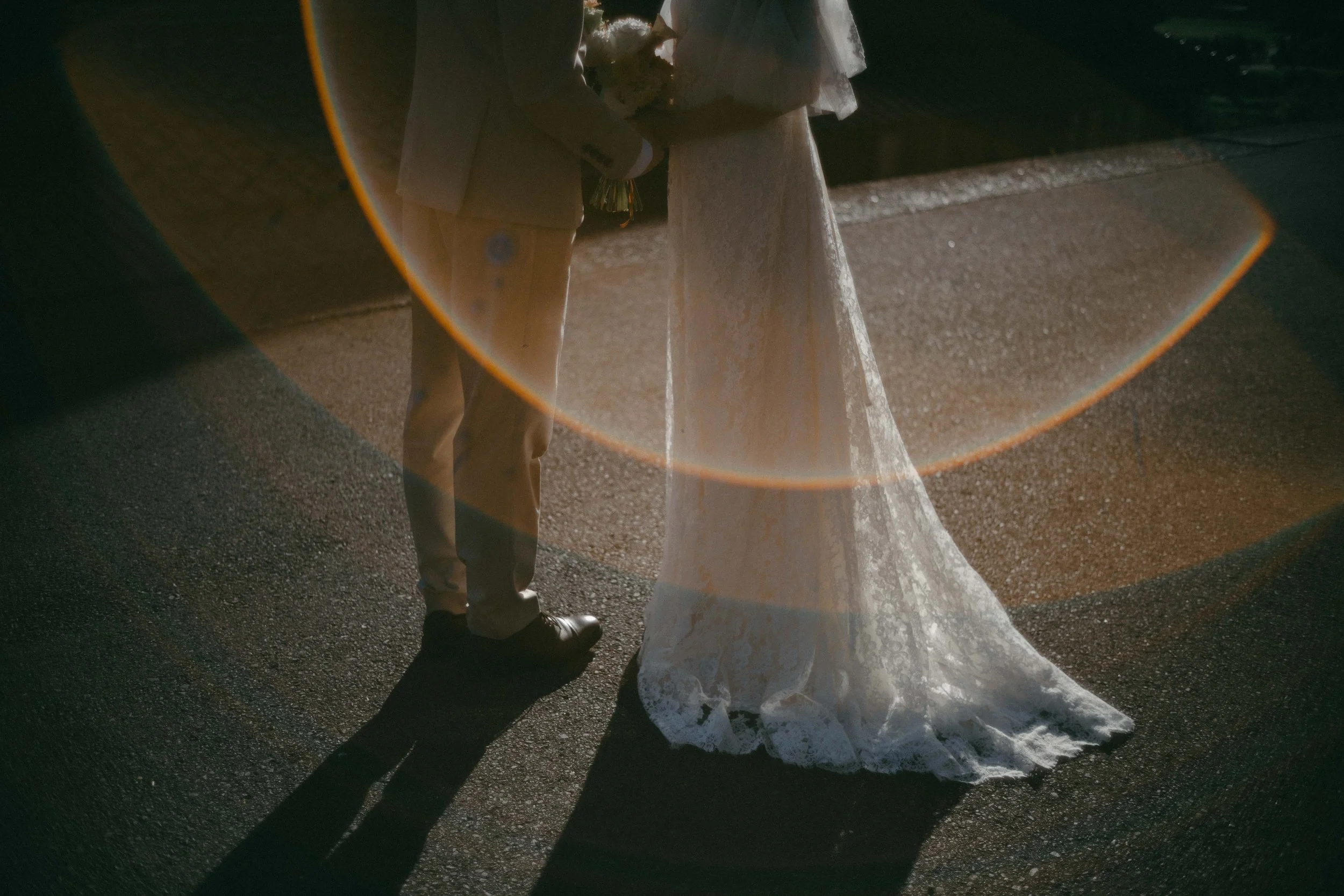 Reflection of a bride in a wedding dress and a groom in a suit holding hands, seen on a car's shiny hood, with sunlight creating a lens flare.