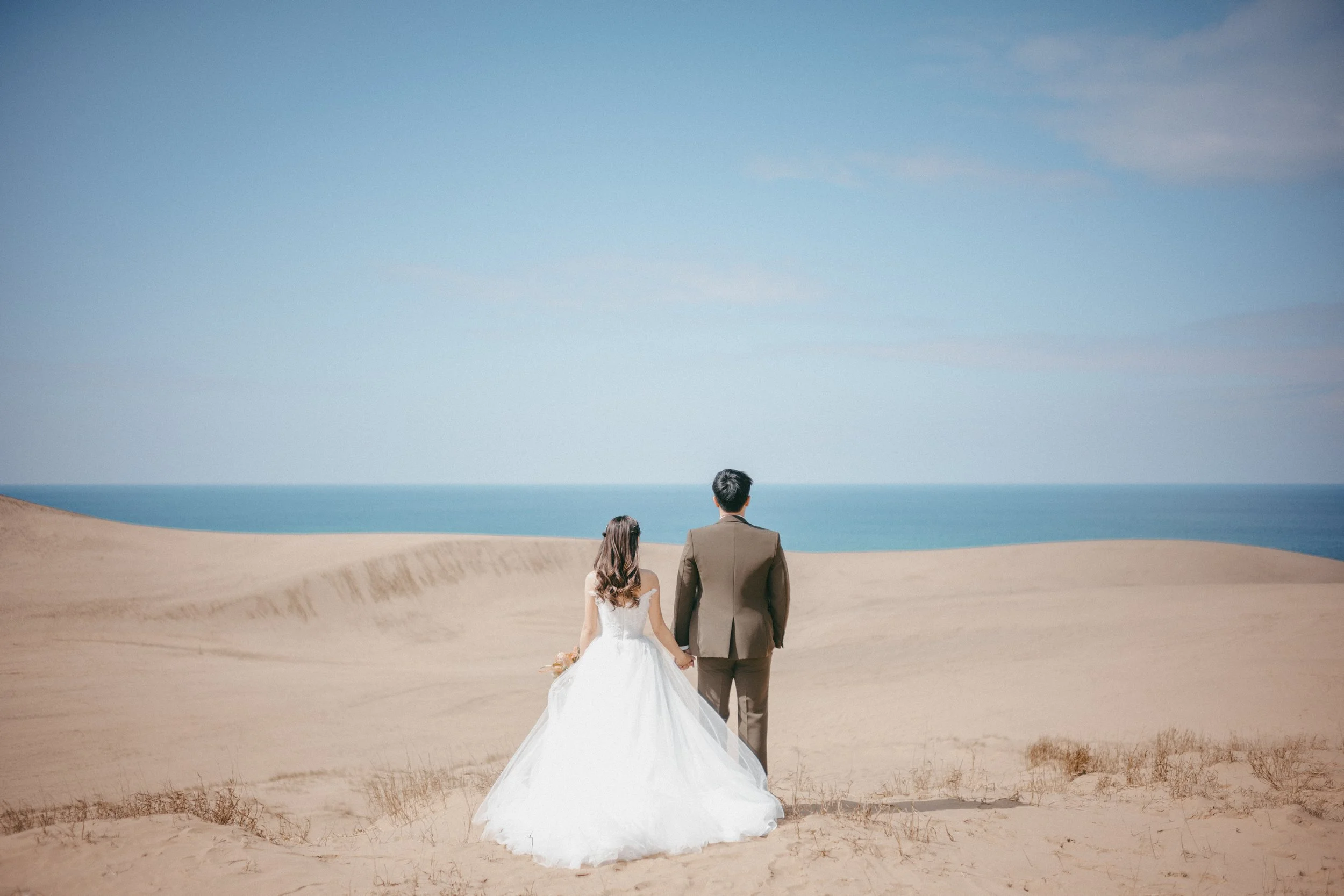A bride in a white wedding gown and a groom in a brown suit holding hands while walking across a sandy desert towards the ocean under a blue sky.