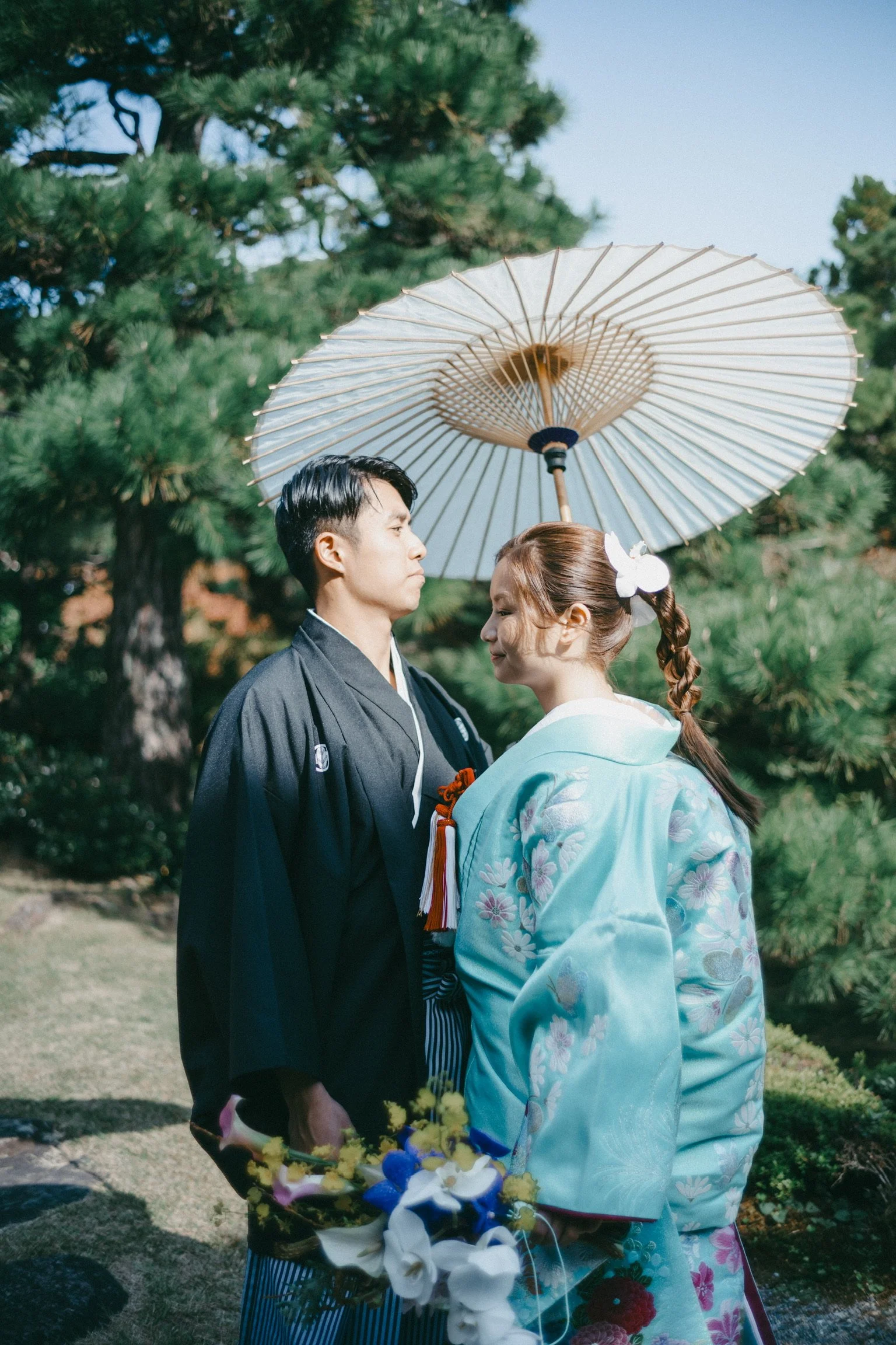 A couple in traditional Japanese attire standing outdoors with a parasol overhead, surrounded by green trees, and the woman holding a bouquet of flowers.