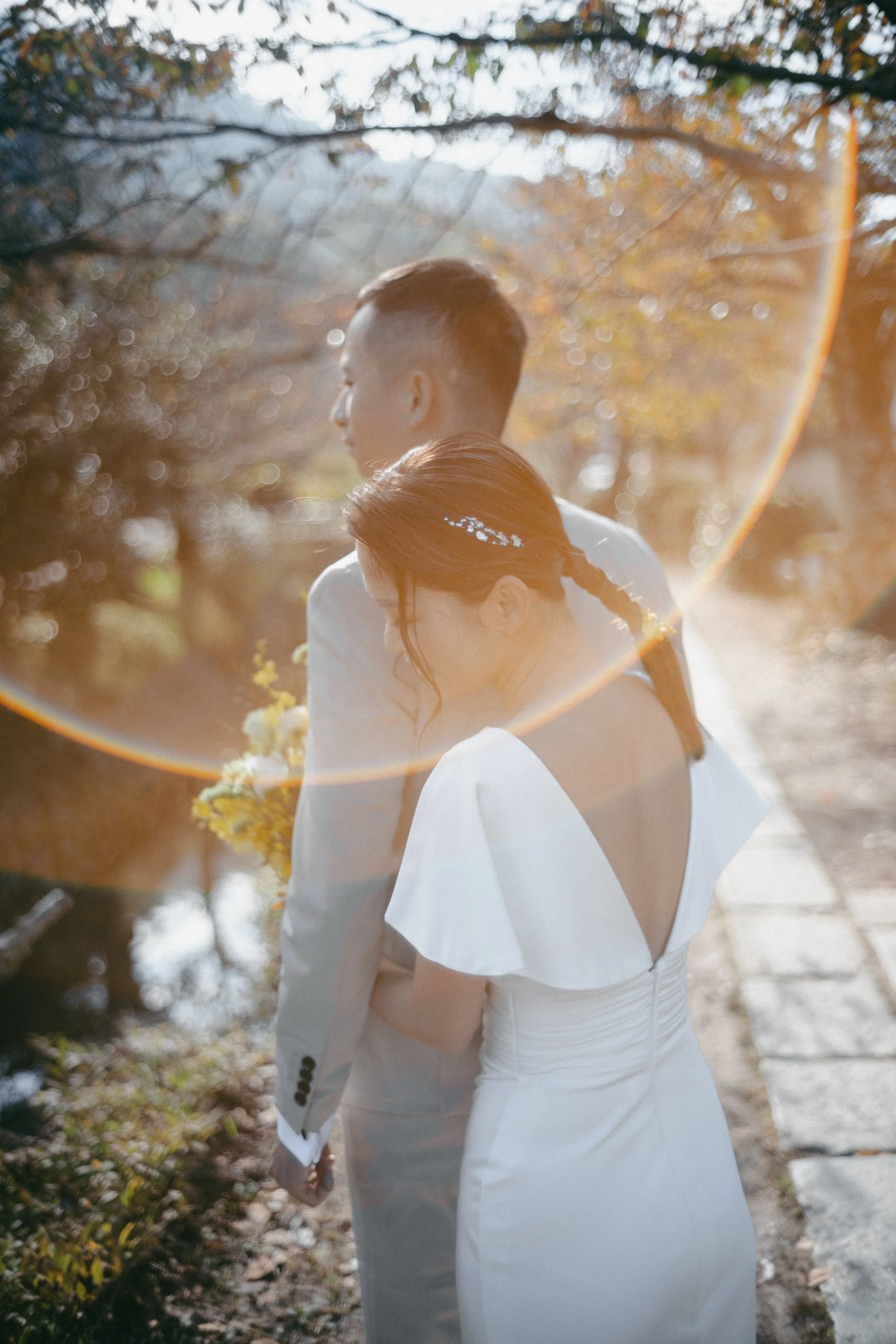 A bride and groom standing close together outdoors in natural light, with a warm, golden glow and lens flare, possibly during sunset, surrounded by trees and a stone path.