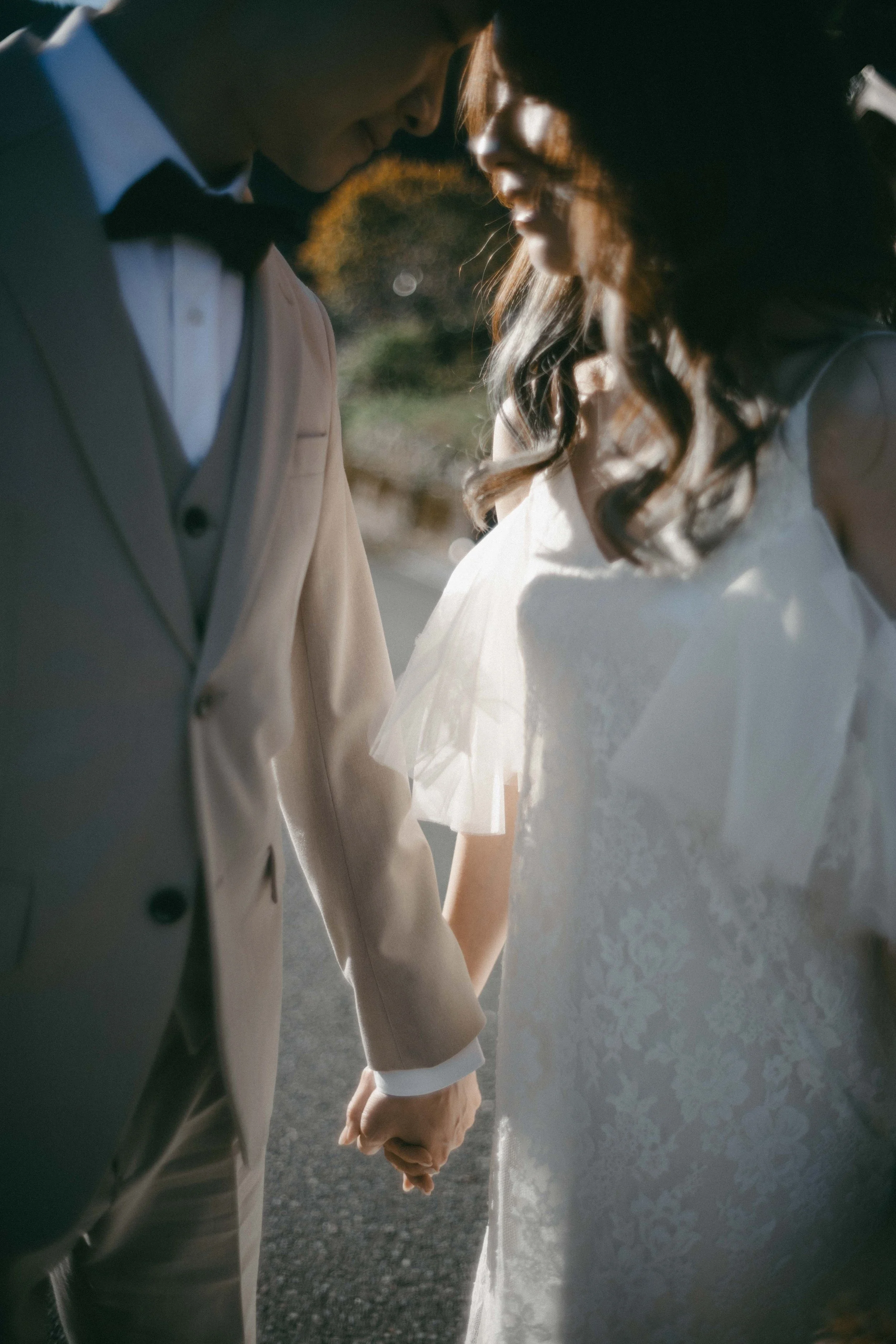 A bride and groom holding hands, dressed in wedding attire, standing close together outdoors during daylight.