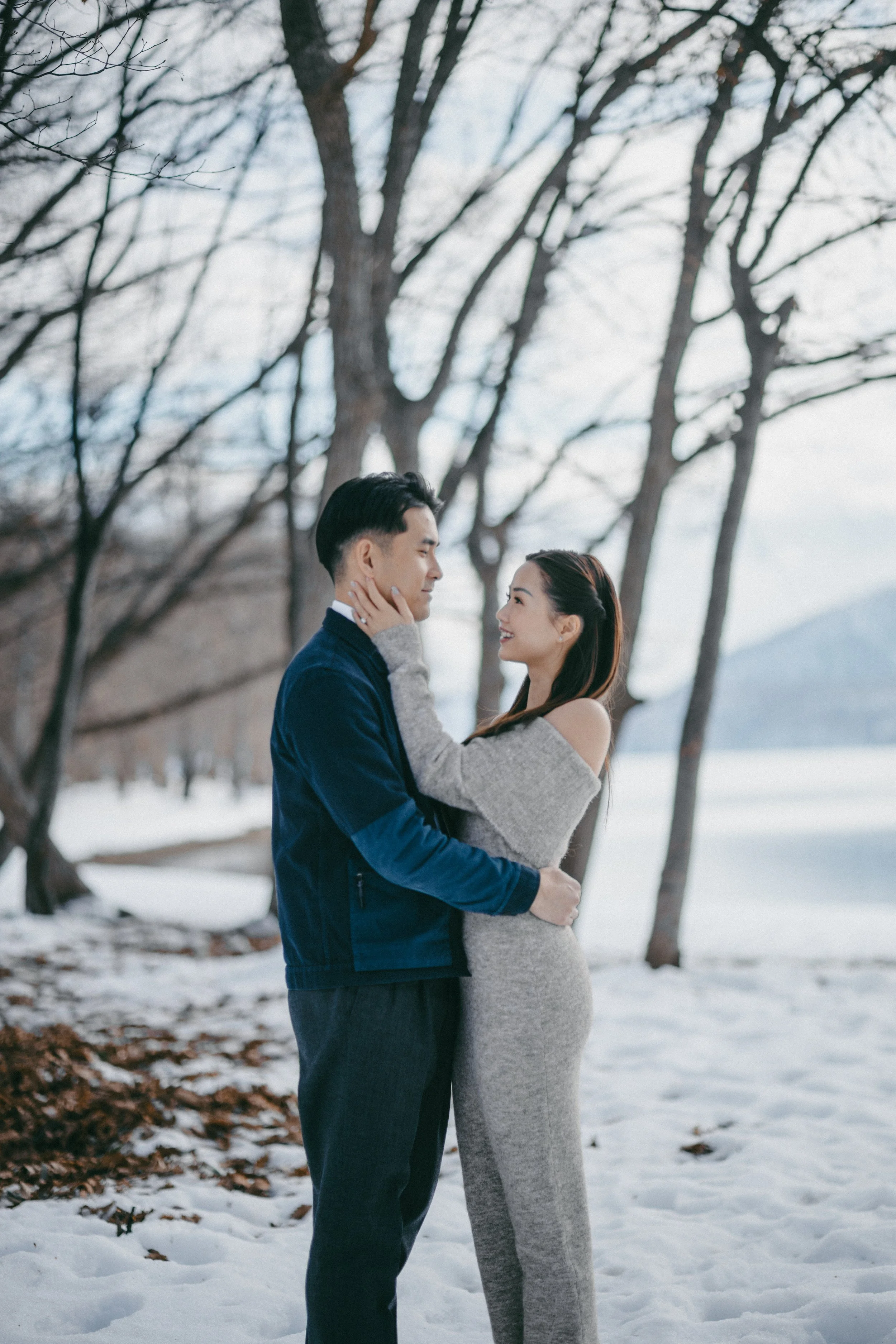 A couple standing closely in a snowy landscape with leafless trees and a frozen lake in the background, smiling at each other.