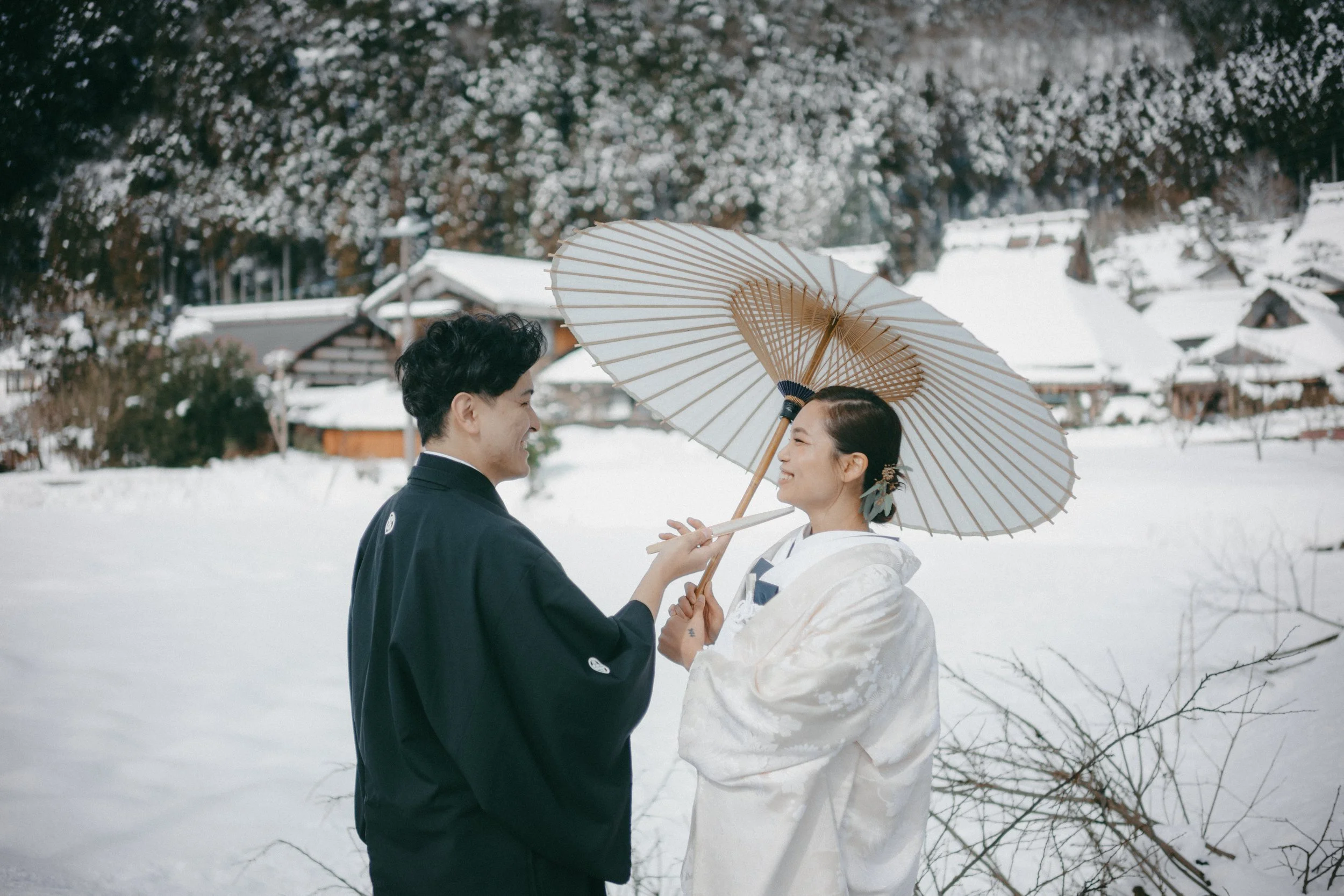 A couple dressed in traditional Japanese wedding attire standing in a snowy landscape, sharing a moment under a white parasol.