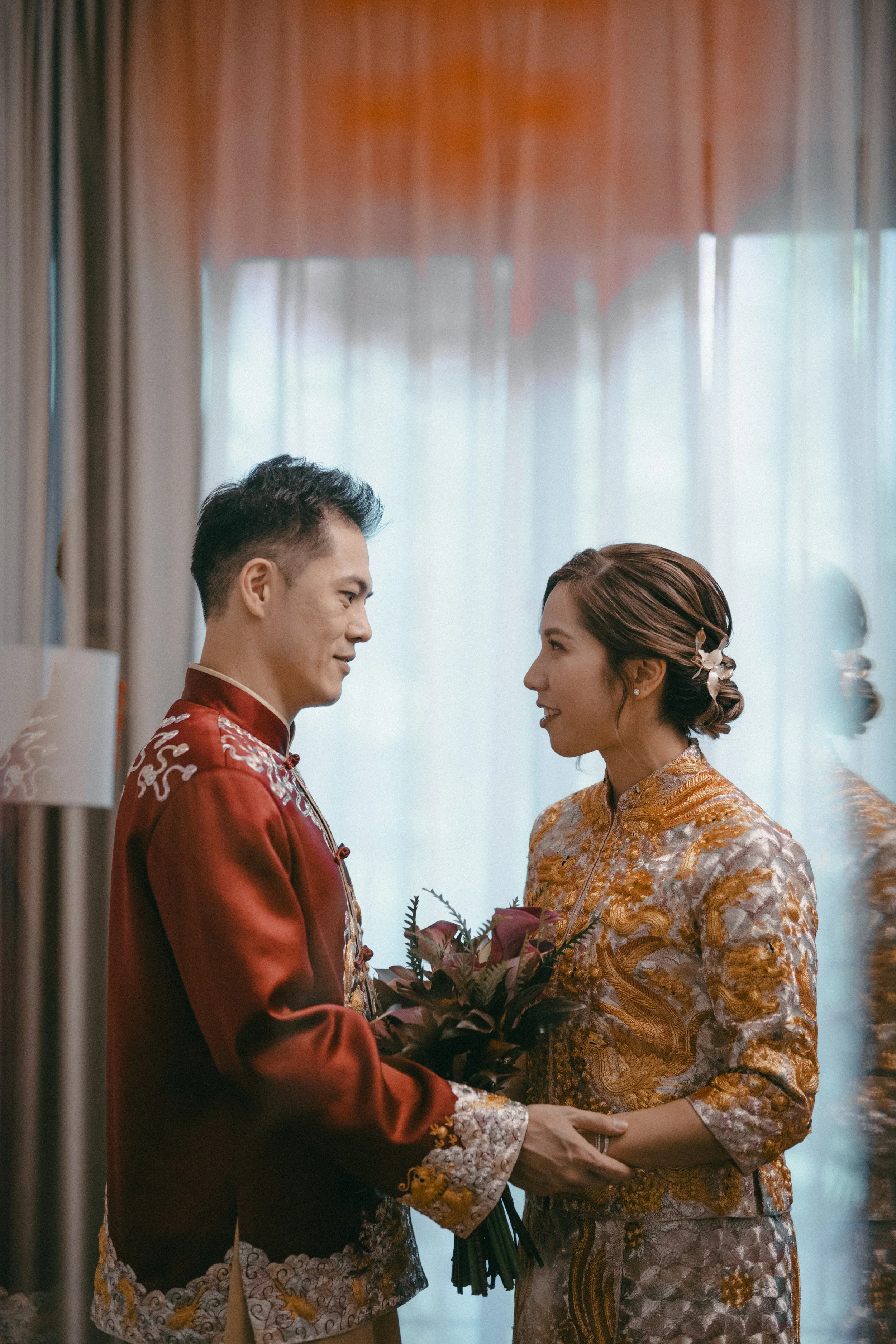 A couple dressed in traditional Chinese wedding attire exchanging a bouquet in a softly lit room.