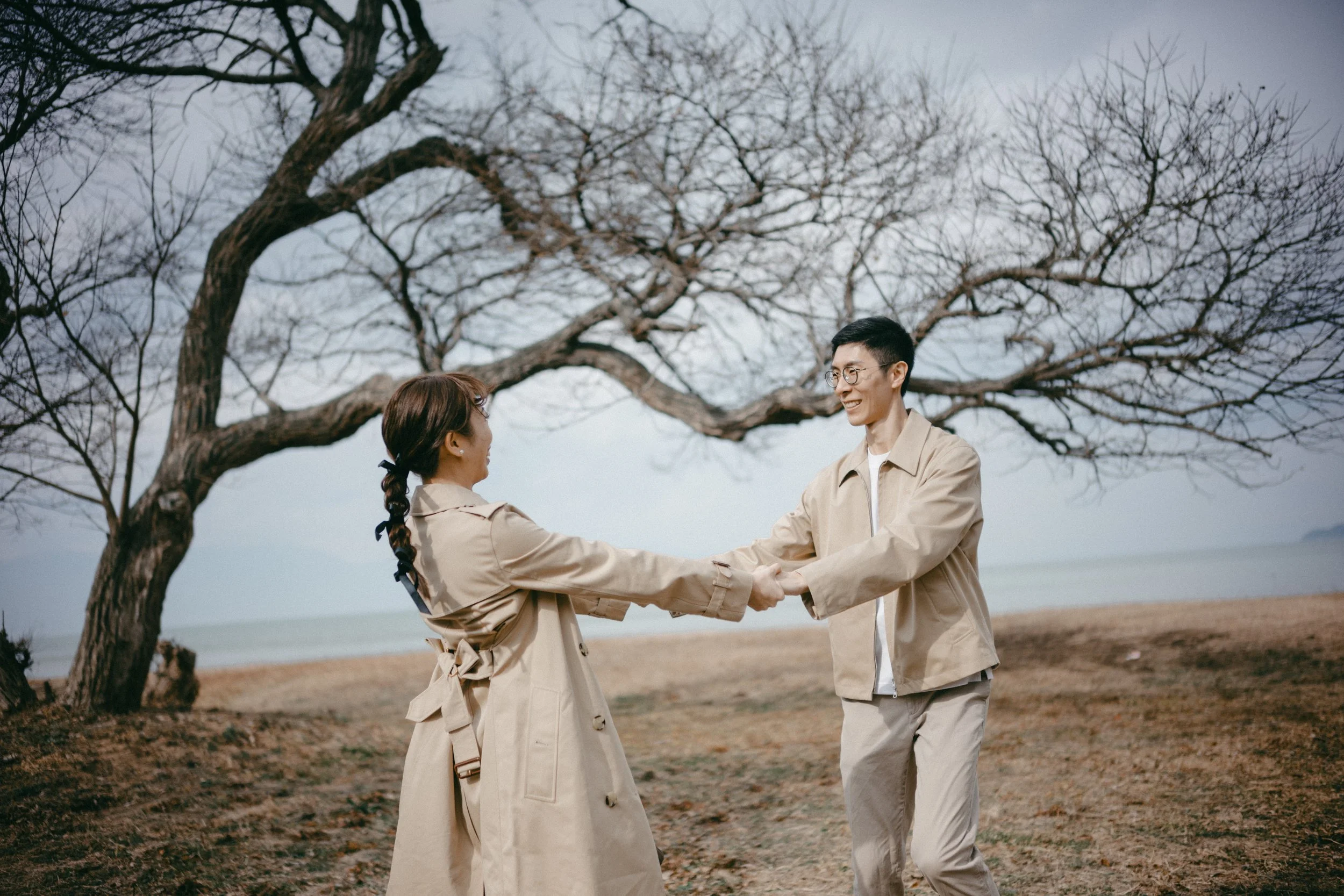 A man and woman in beige coats holding hands and smiling on a dirt path near a leafless tree with the ocean in the background.