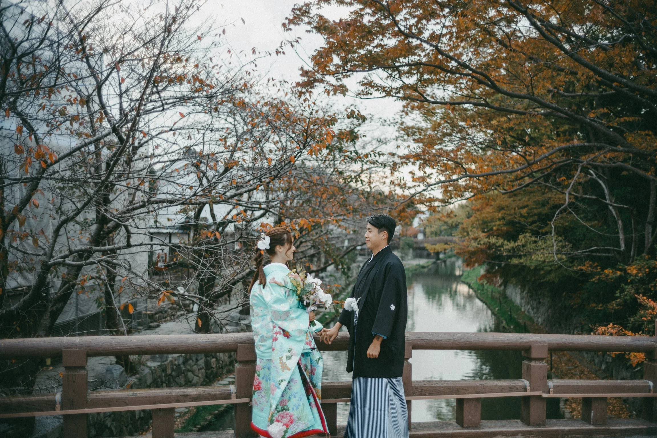 A couple in traditional Japanese wedding attire holding hands on a bridge over a river surrounded by autumn trees.