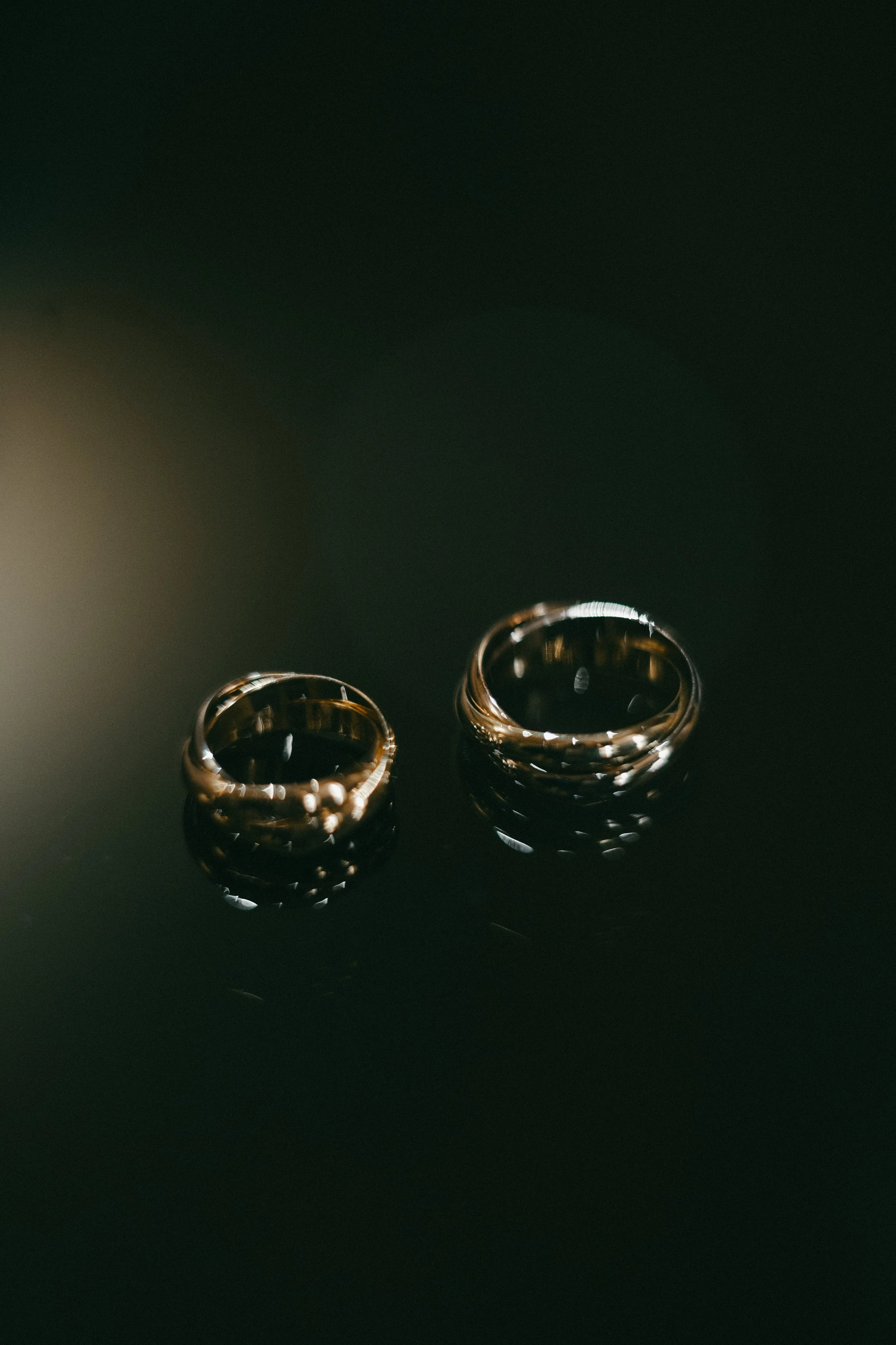 Two gold wedding bands, one smaller and one larger, placed on a dark surface with soft lighting.