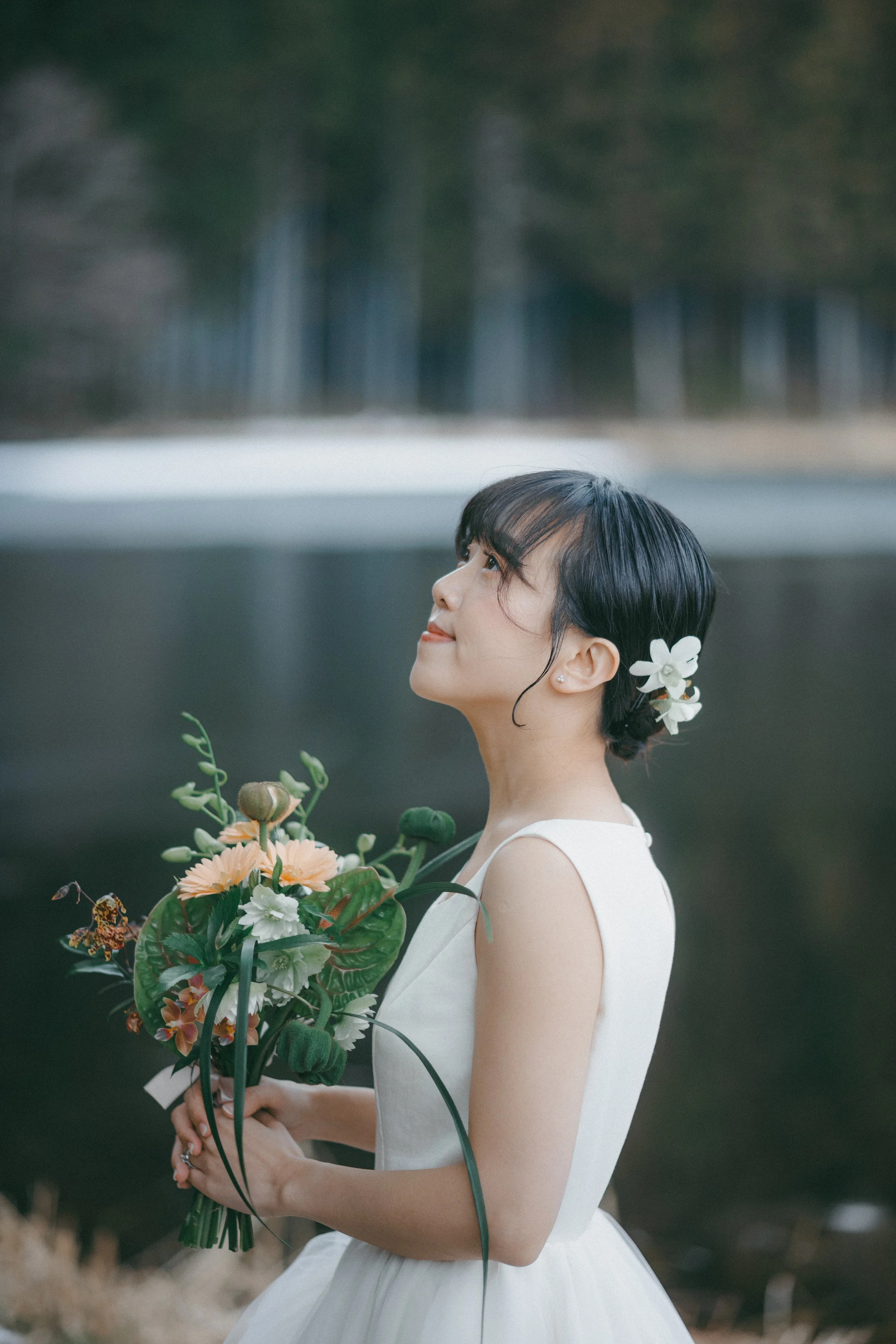 A woman in a white dress holding a bouquet of flowers, standing outdoors near a body of water with trees in the background.