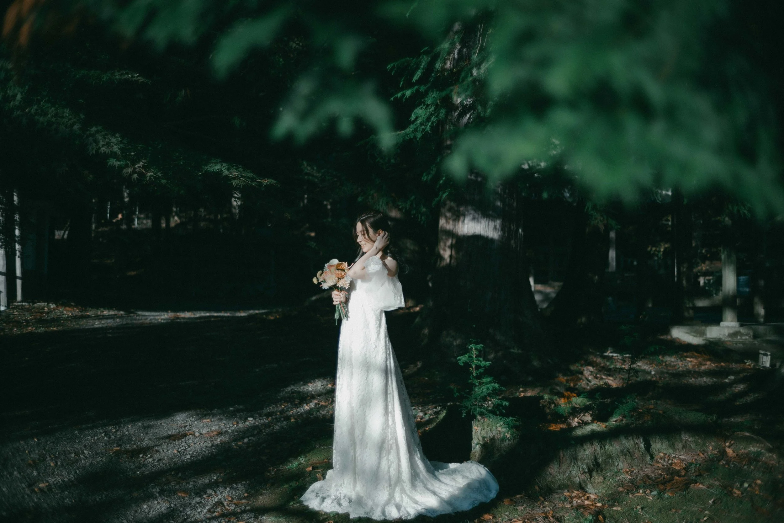 A woman in a white wedding dress holding a bouquet of flowers standing outdoors under trees.