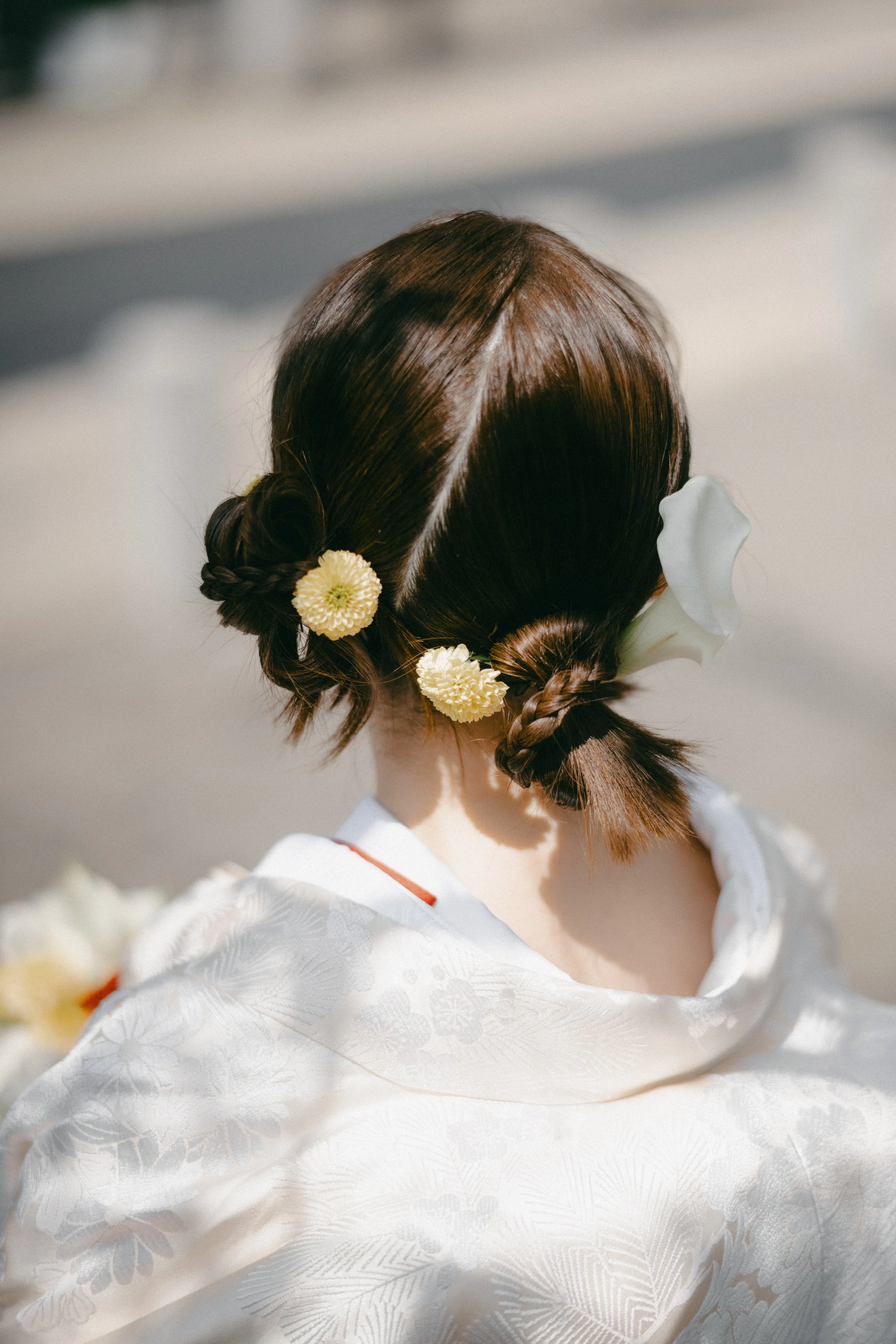 Back of a woman's head with traditional Japanese hairstyle decorated with white and yellow flowers, wearing a white kimono with intricate floral patterns.