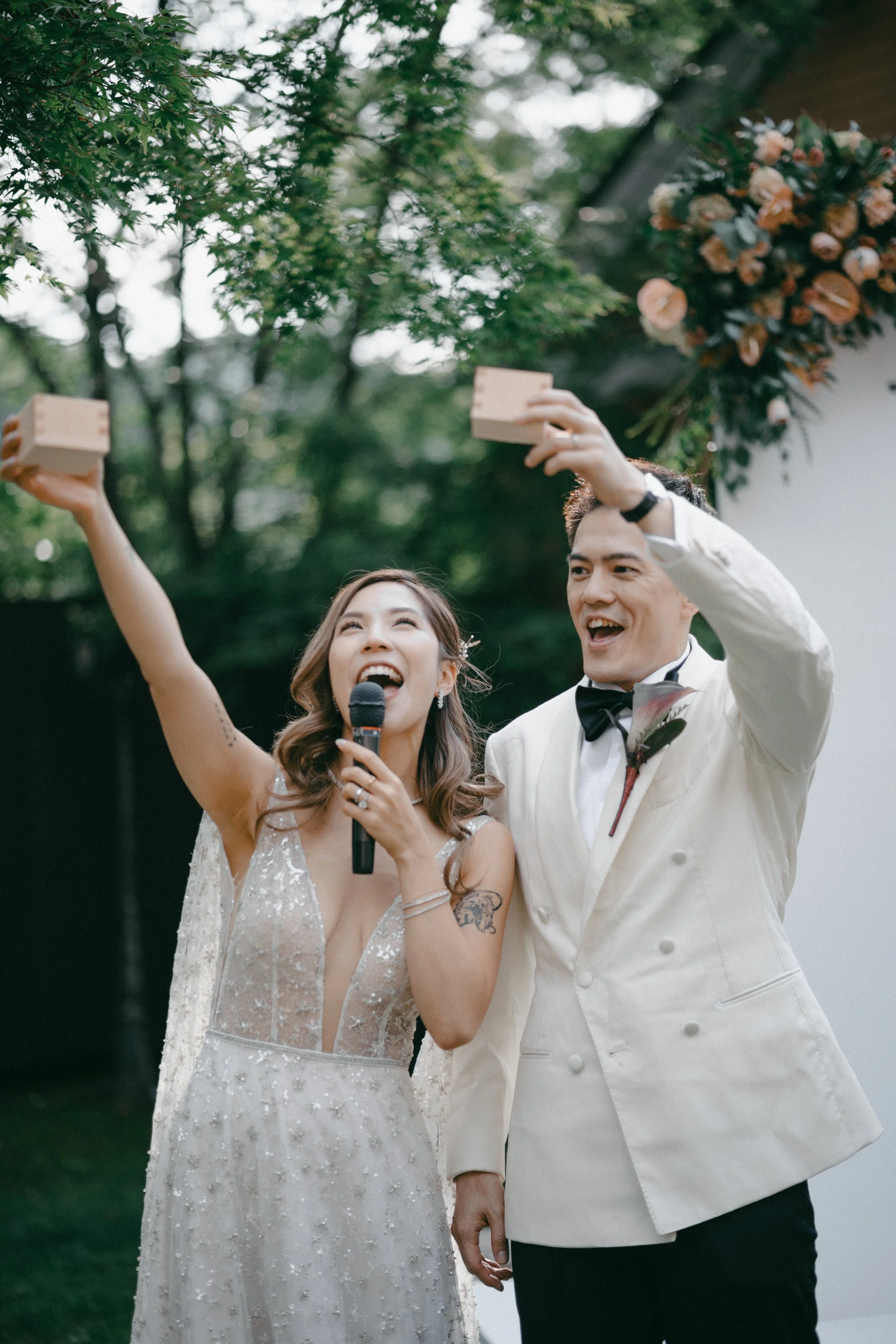 A bride and groom at their wedding ceremony, smiling and raising their arms, with the bride holding a microphone and the groom holding a small wooden block, outdoors with greenery and floral arrangements in the background.