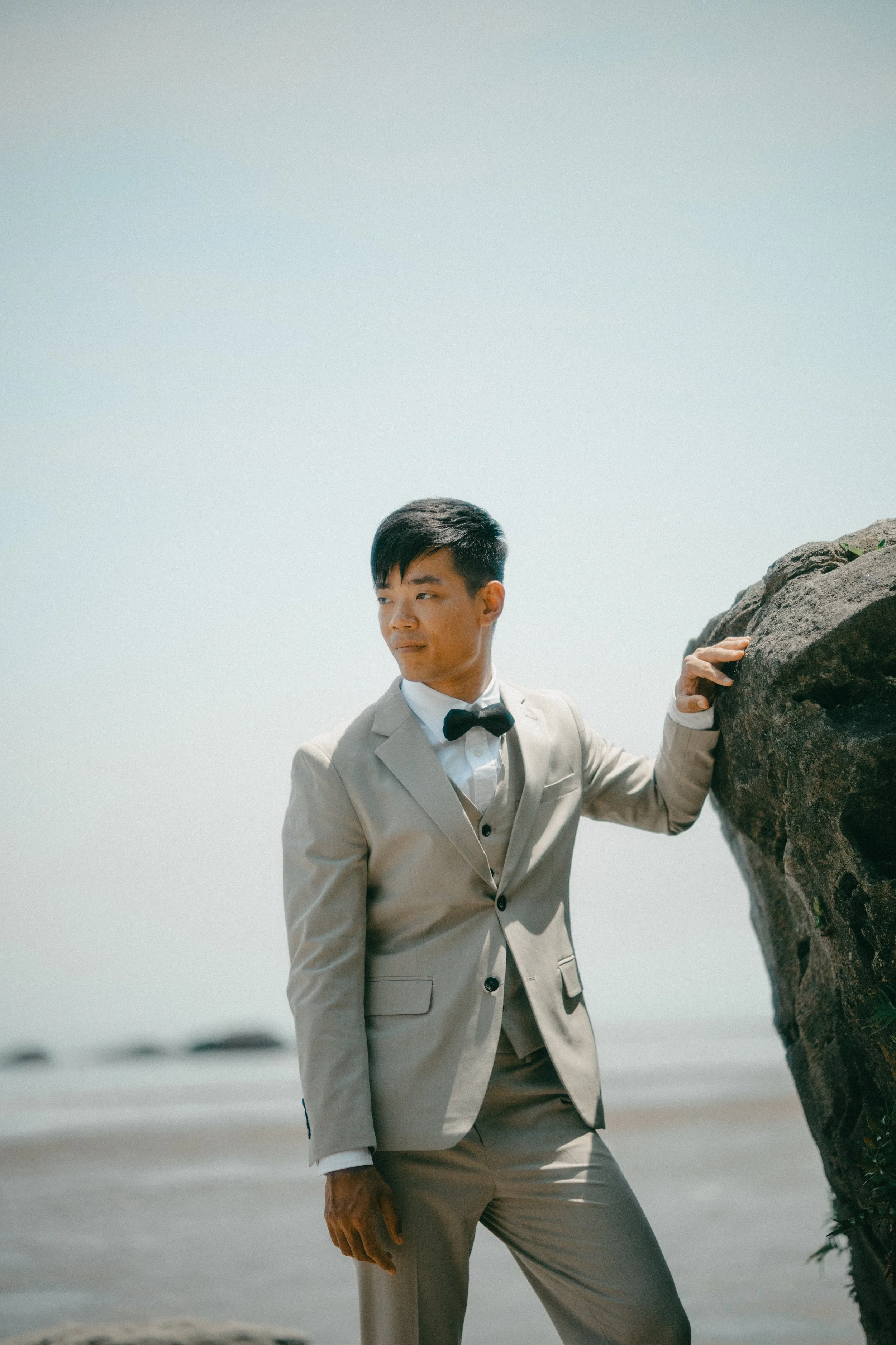 A man in a beige suit and black bow tie standing on a beach, leaning on a large rock with the ocean in the background.