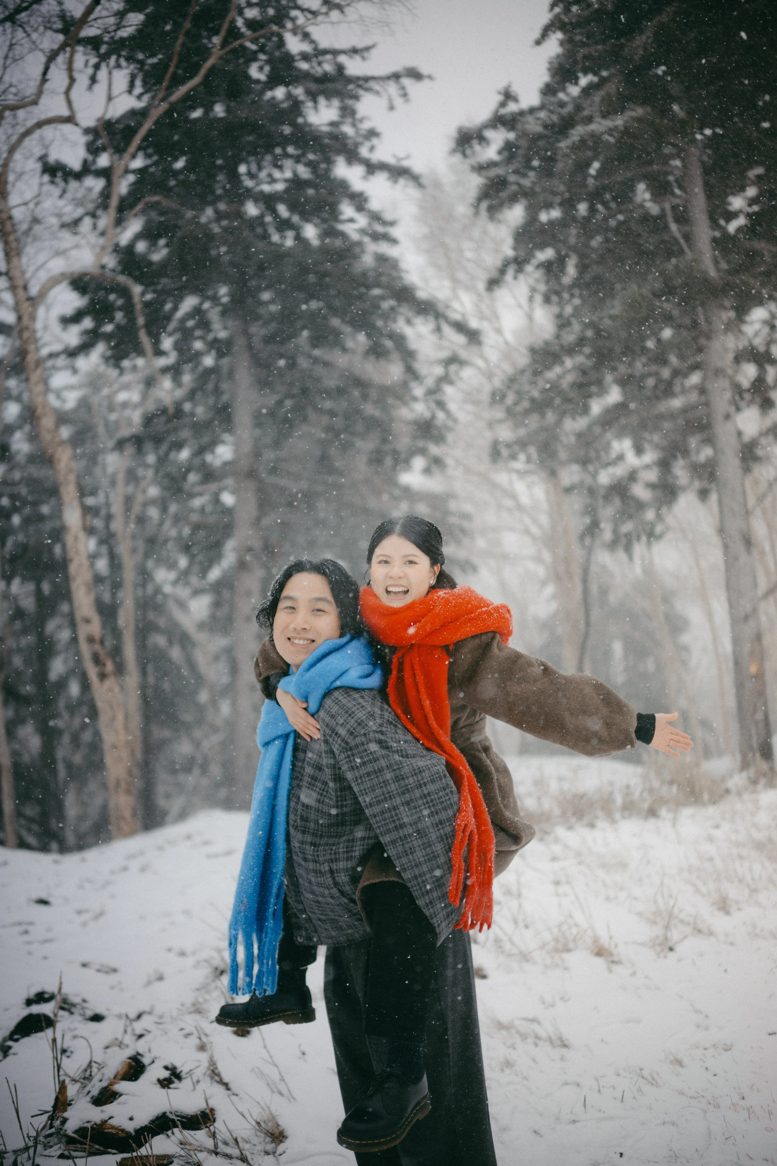 A man giving a woman a piggyback ride in a snowy forest, both smiling and wearing winter clothes and scarves.