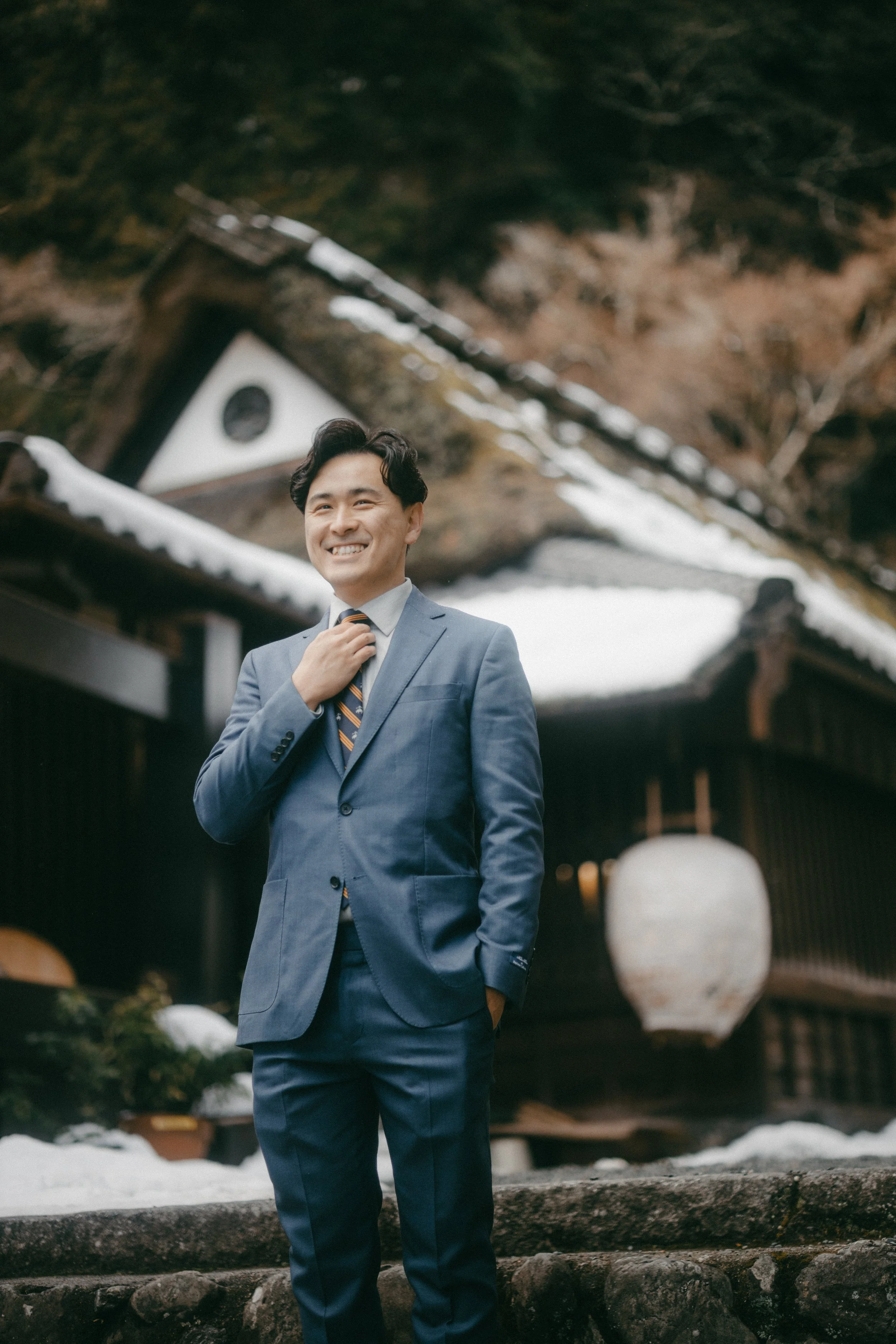 A young man in a gray suit and striped tie stands outdoors, smiling confidently, with traditional Japanese buildings and snow in the background.