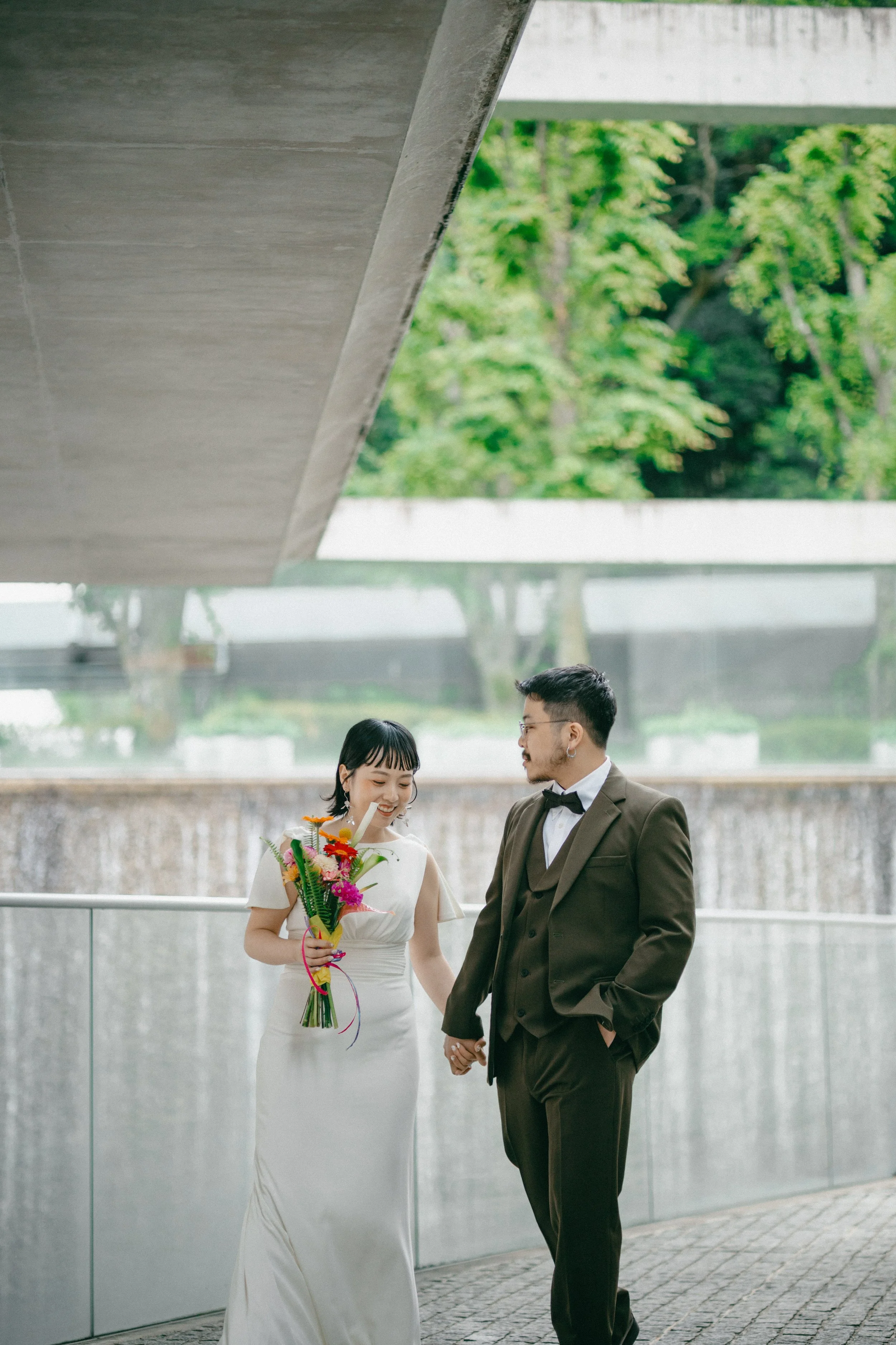 A couple dressed in wedding attire holding hands outdoors with a modern building and green trees in the background.