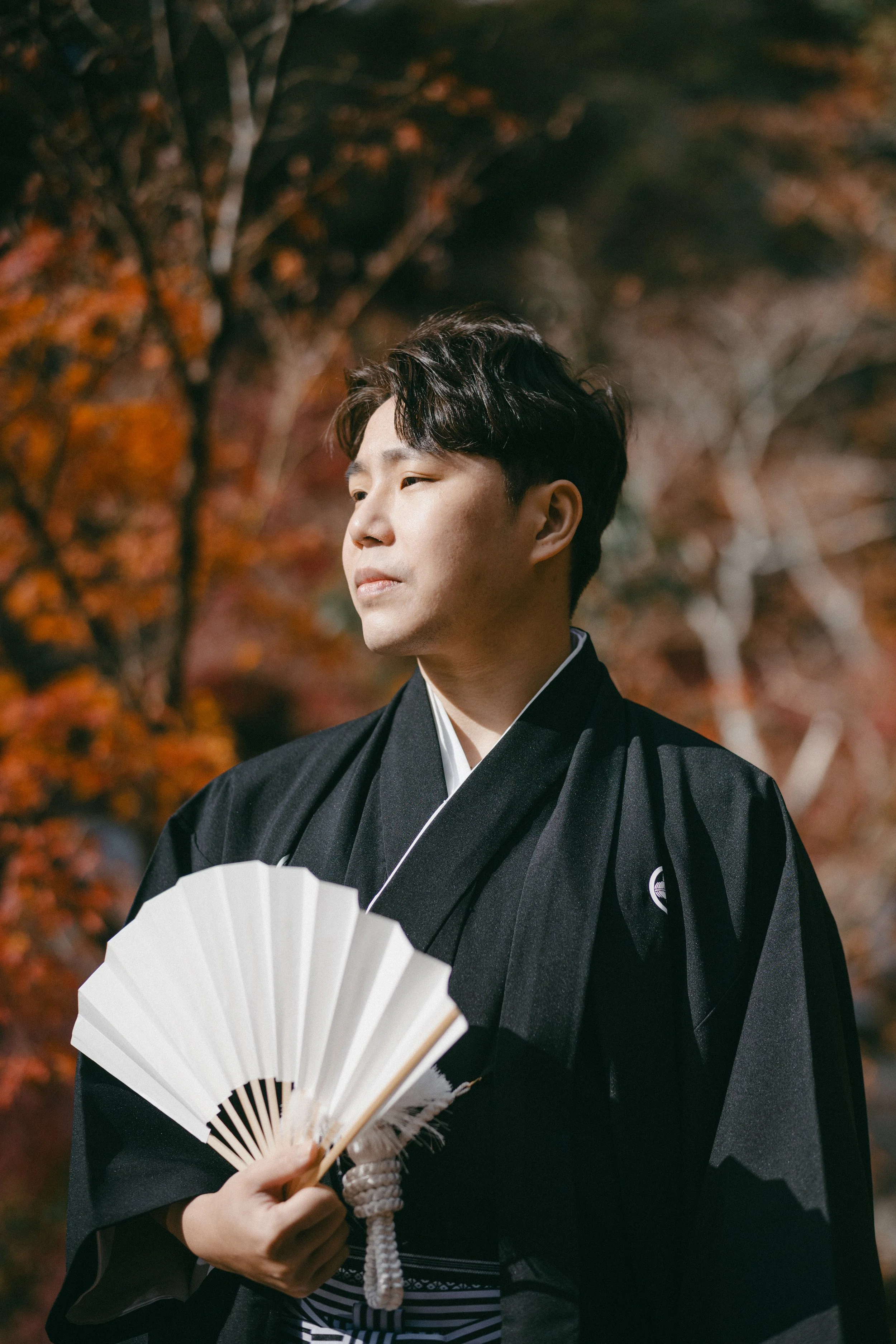 A young man dressed in traditional Japanese attire holding a paper fan, standing outdoors with autumn foliage in the background.