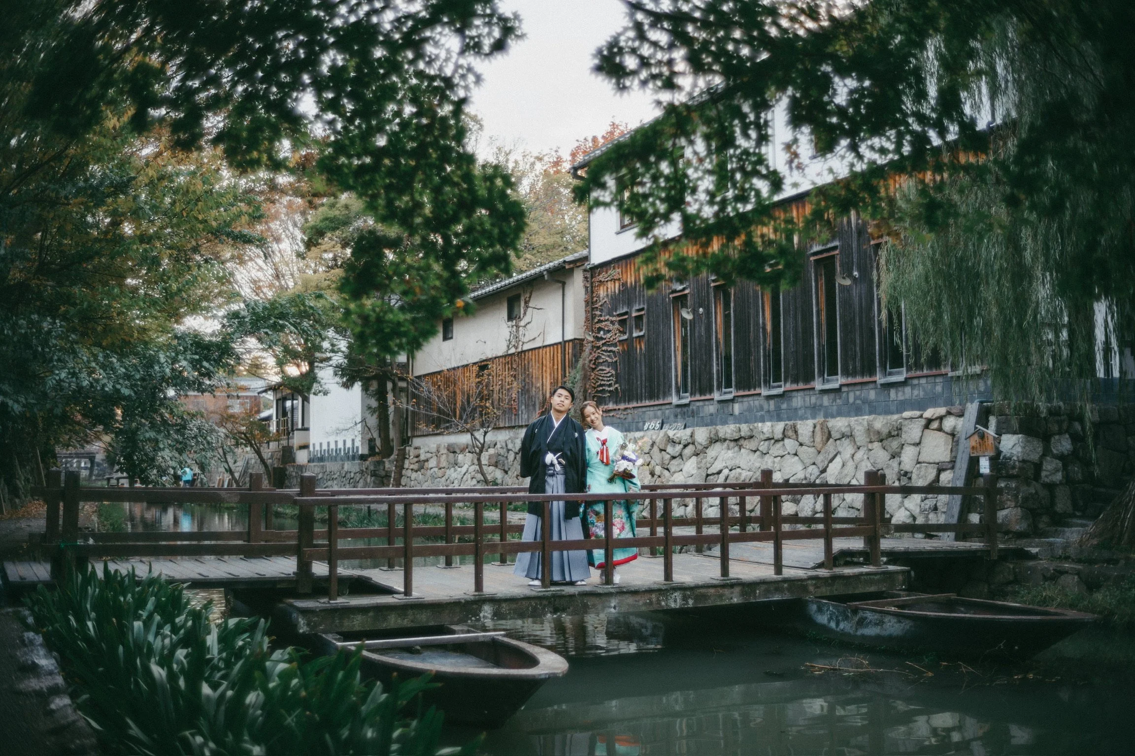 Two people dressed in traditional Japanese kimono and hakama standing on a small wooden bridge over a pond, with autumn trees and traditional buildings in the background.