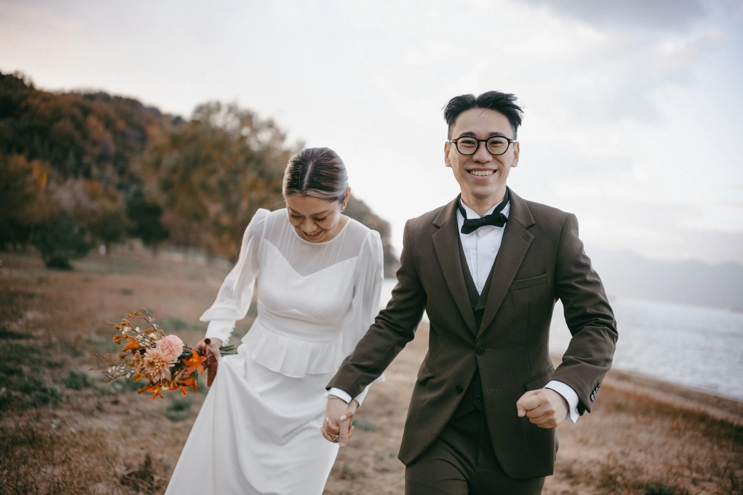 A smiling man in a tuxedo holding hands with a woman in a white wedding dress holding a bouquet, walking along a beach with trees and water in the background.
