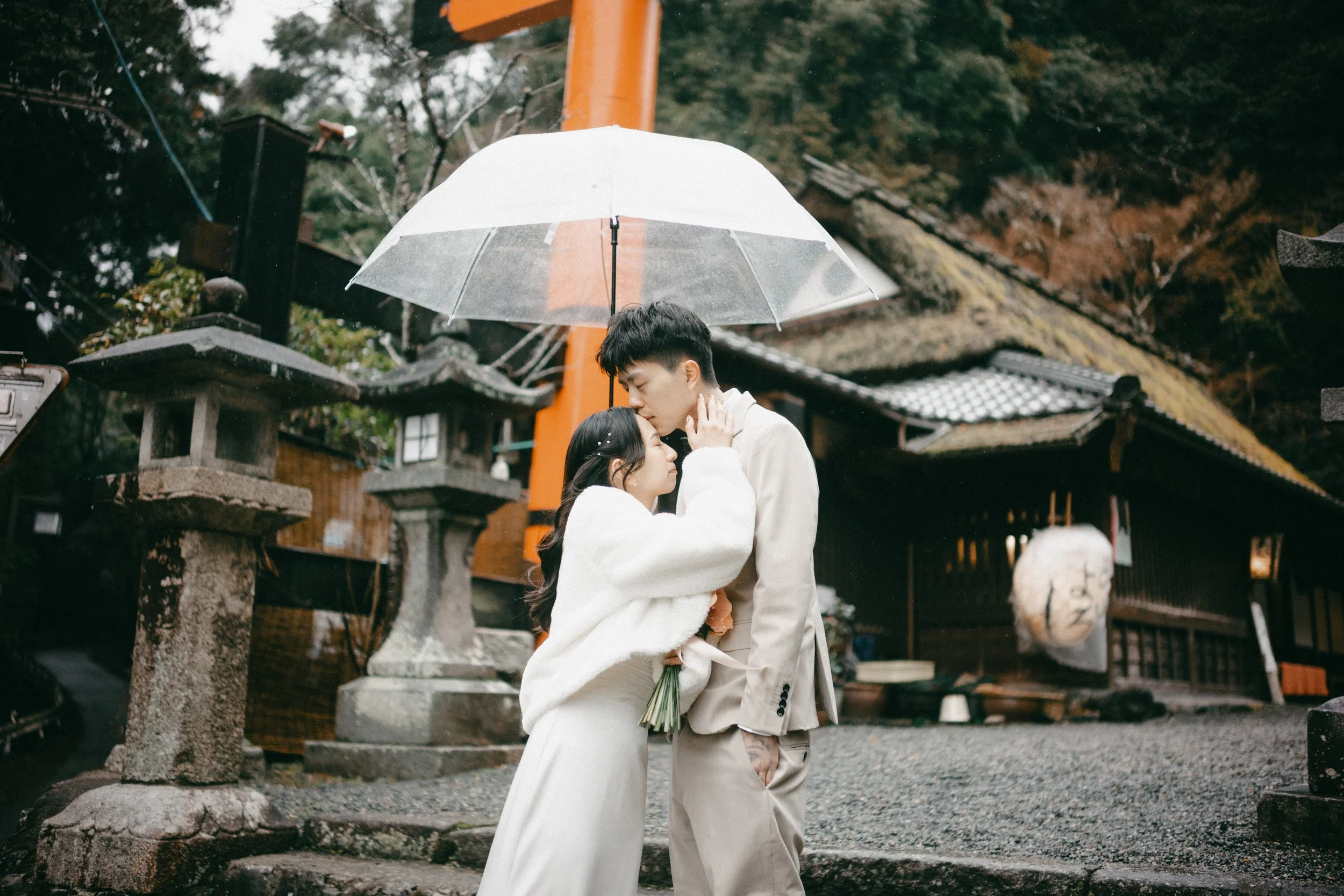 A couple standing close together under an umbrella in a traditional Japanese outdoor setting on a rainy day. The woman is holding flowers, and both are dressed in light-colored clothing. The background features stone lanterns, a wooden building, and 