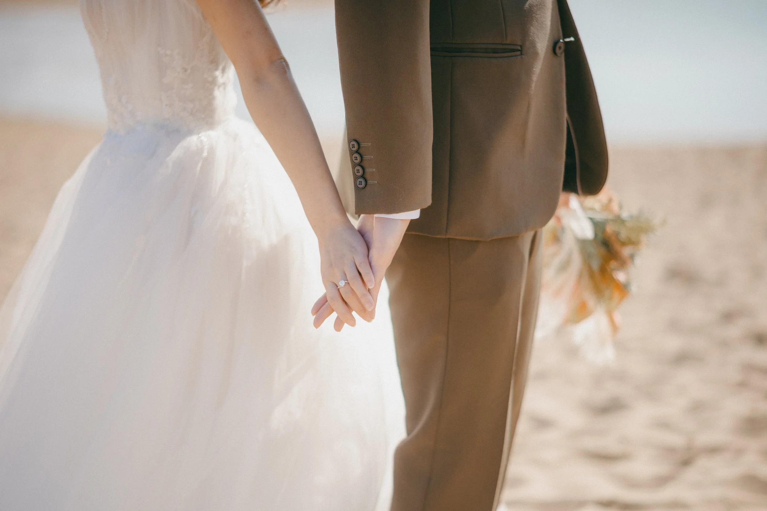 A bride and groom holding hands on the beach with wedding rings, the bride in a white gown and the groom in a brown suit.