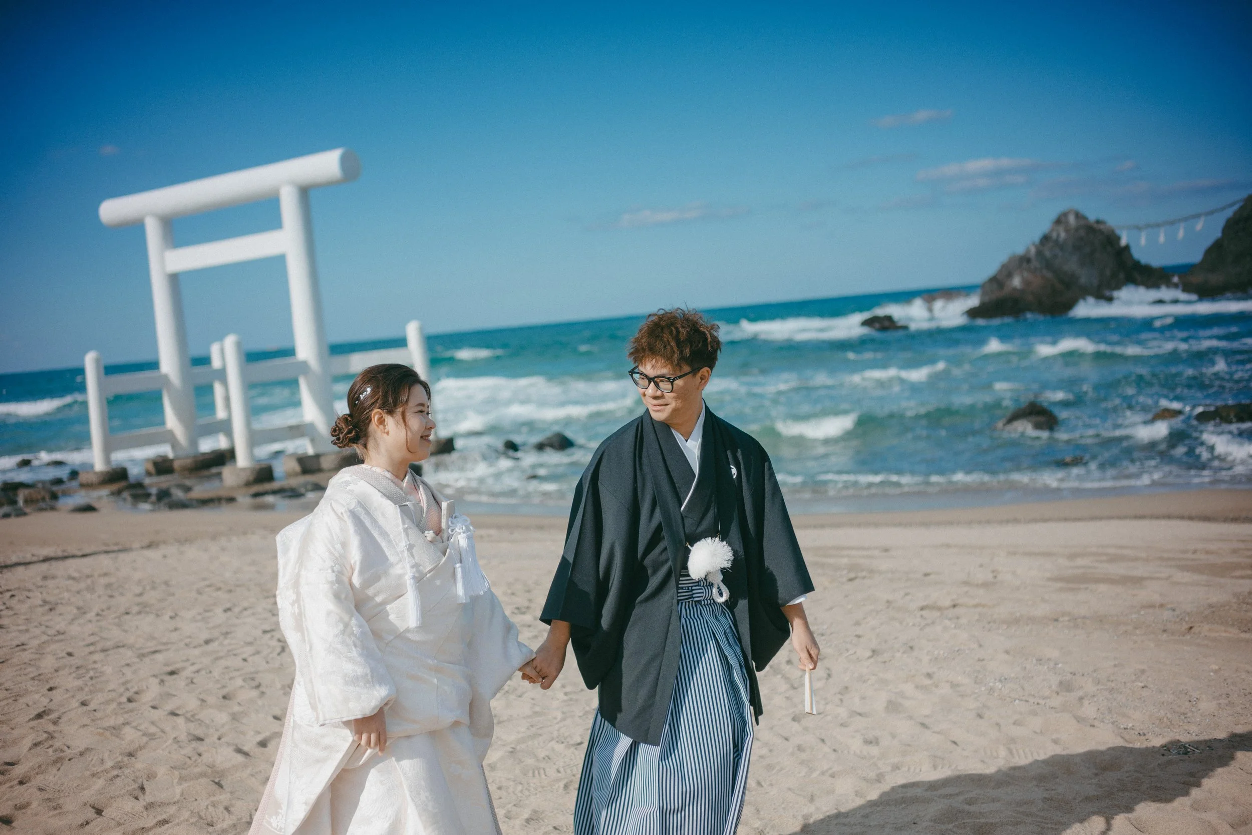 A couple dressed in traditional Japanese wedding attire walking on a beach holding hands, with a torii gate and ocean in the background.