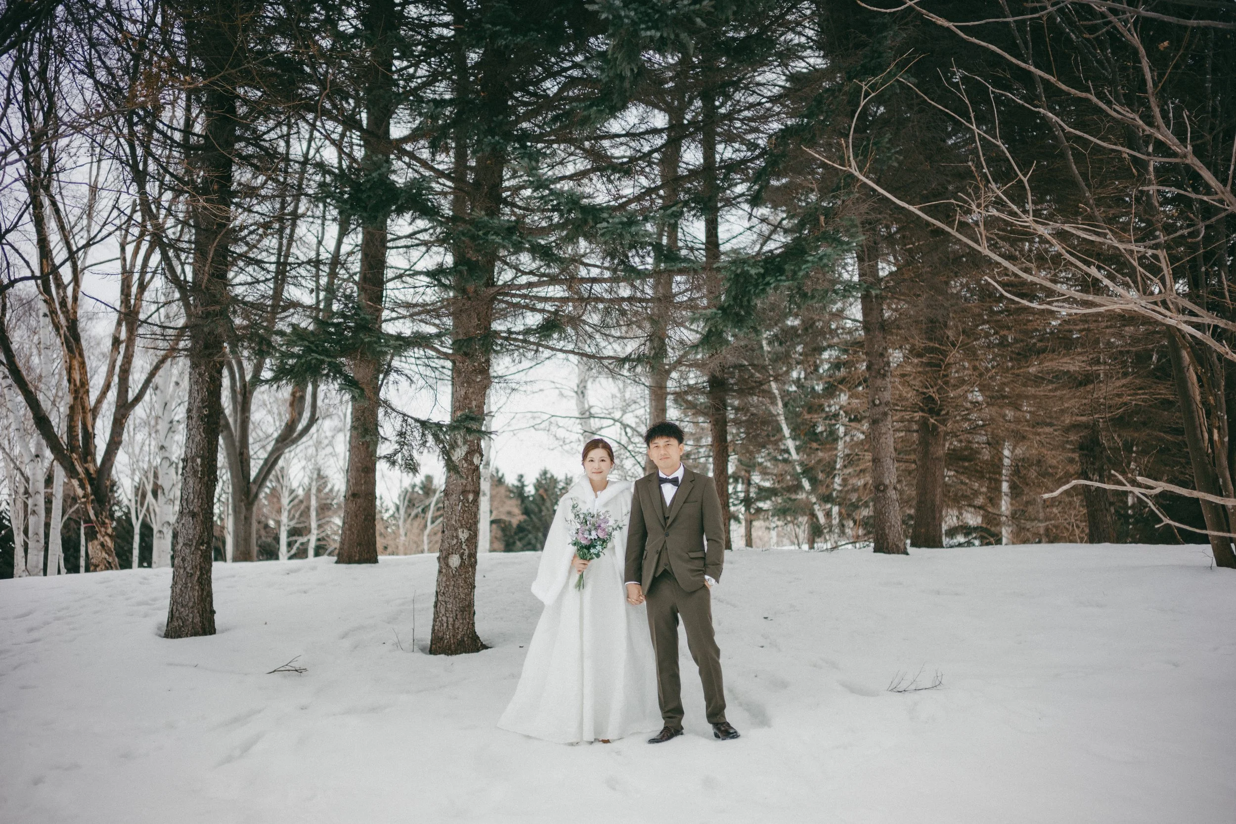 A wedding couple holding hands outdoors in a snowy forest, with the bride in a white gown and veil holding a bouquet, and the groom in a suit with a bowtie, standing beneath tall trees.