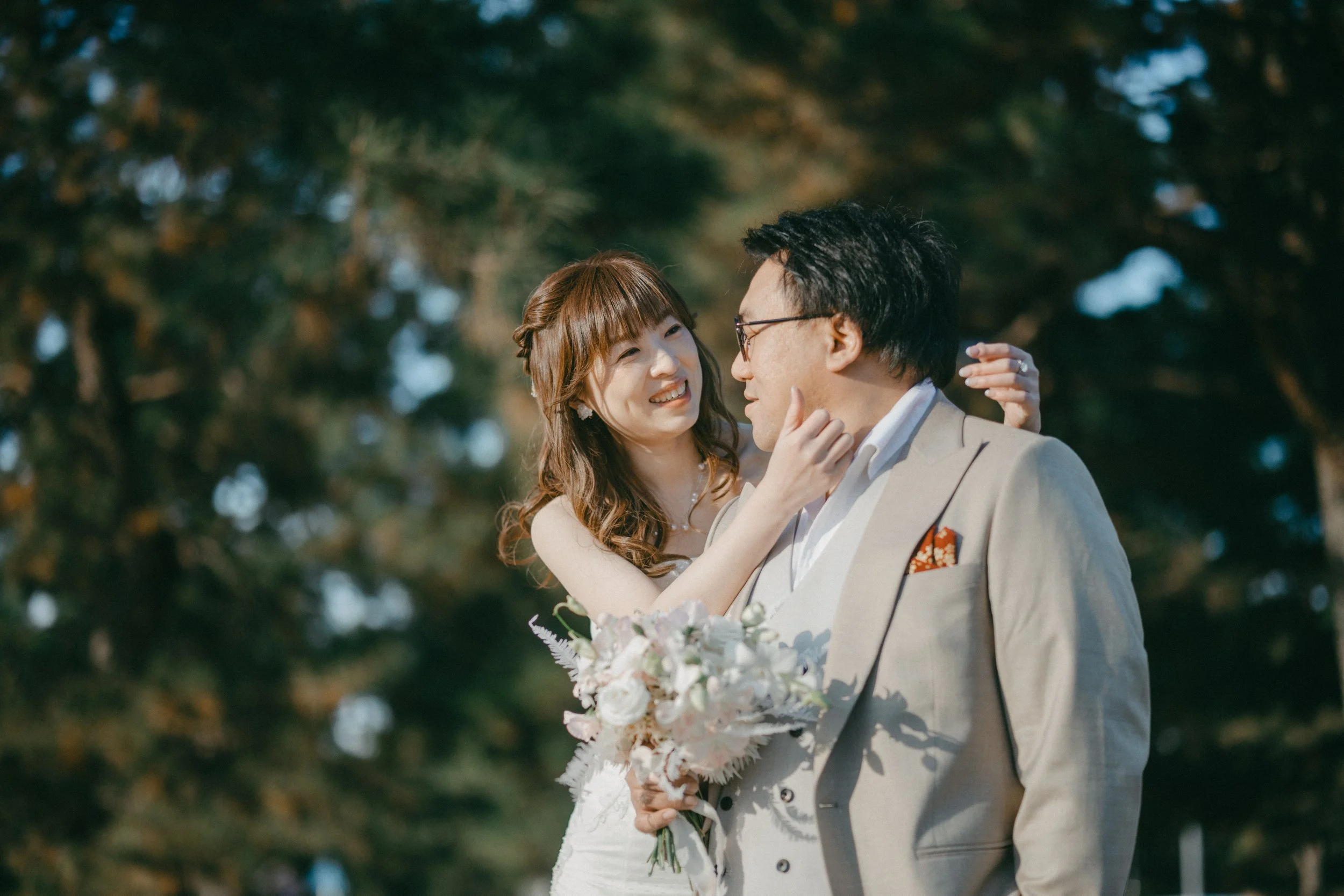 A woman on her wedding day with a man, holding a bouquet of flowers, smiling at each other outdoors surrounded by trees.