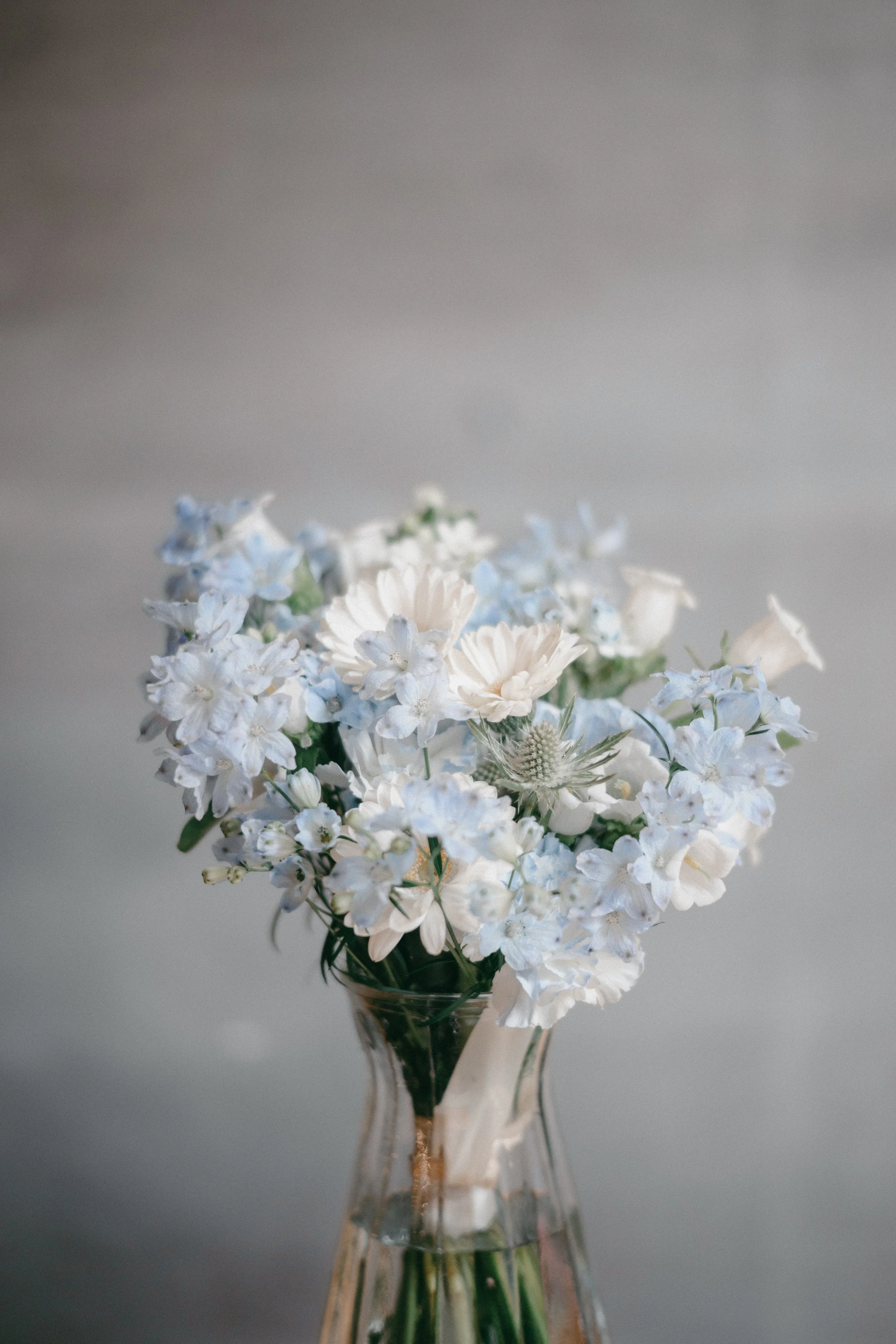 A clear glass vase with a bouquet of white and light blue flowers against a neutral background.