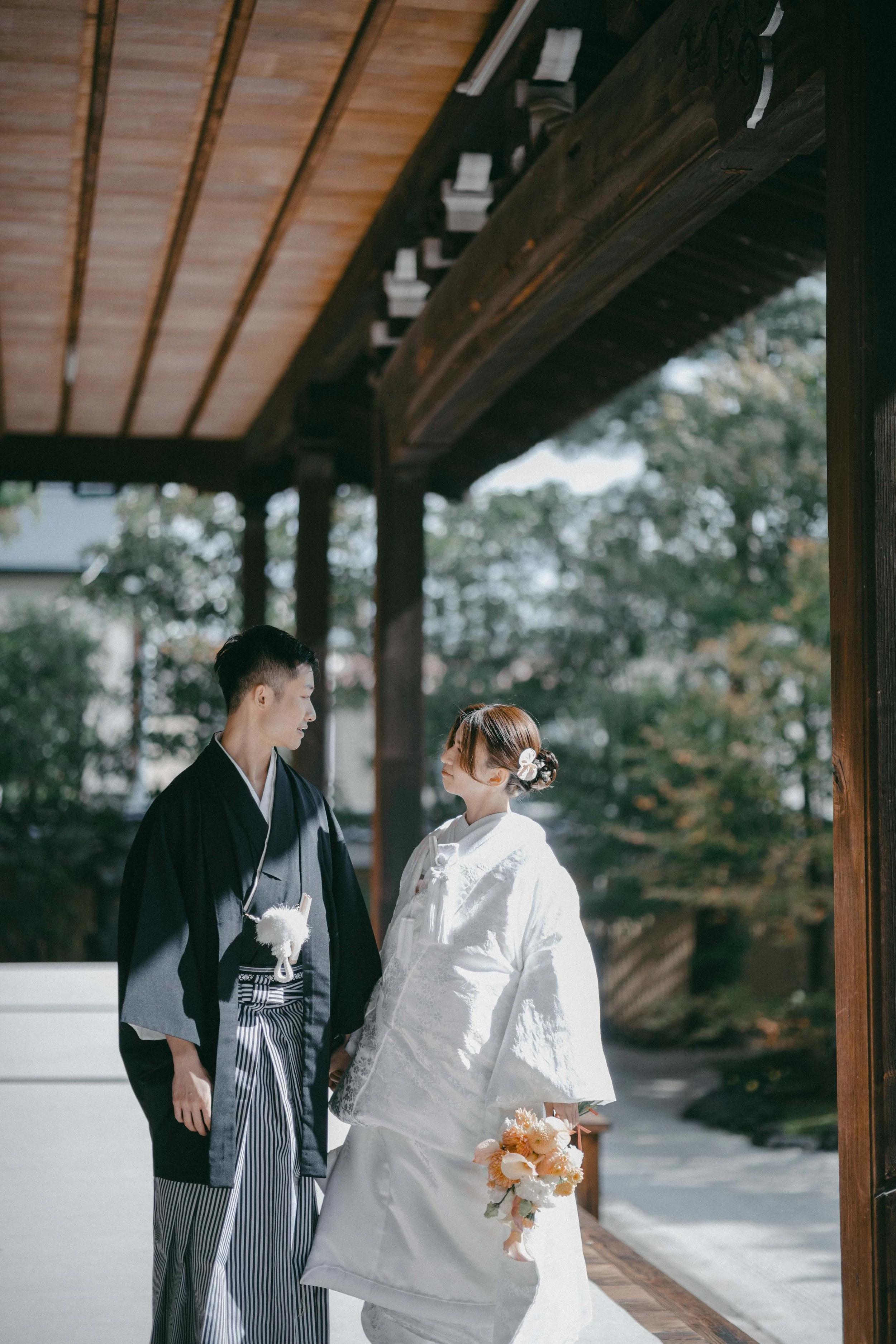 A couple dressed in traditional Japanese wedding attire standing under a wooden structure outdoors, with trees in the background.