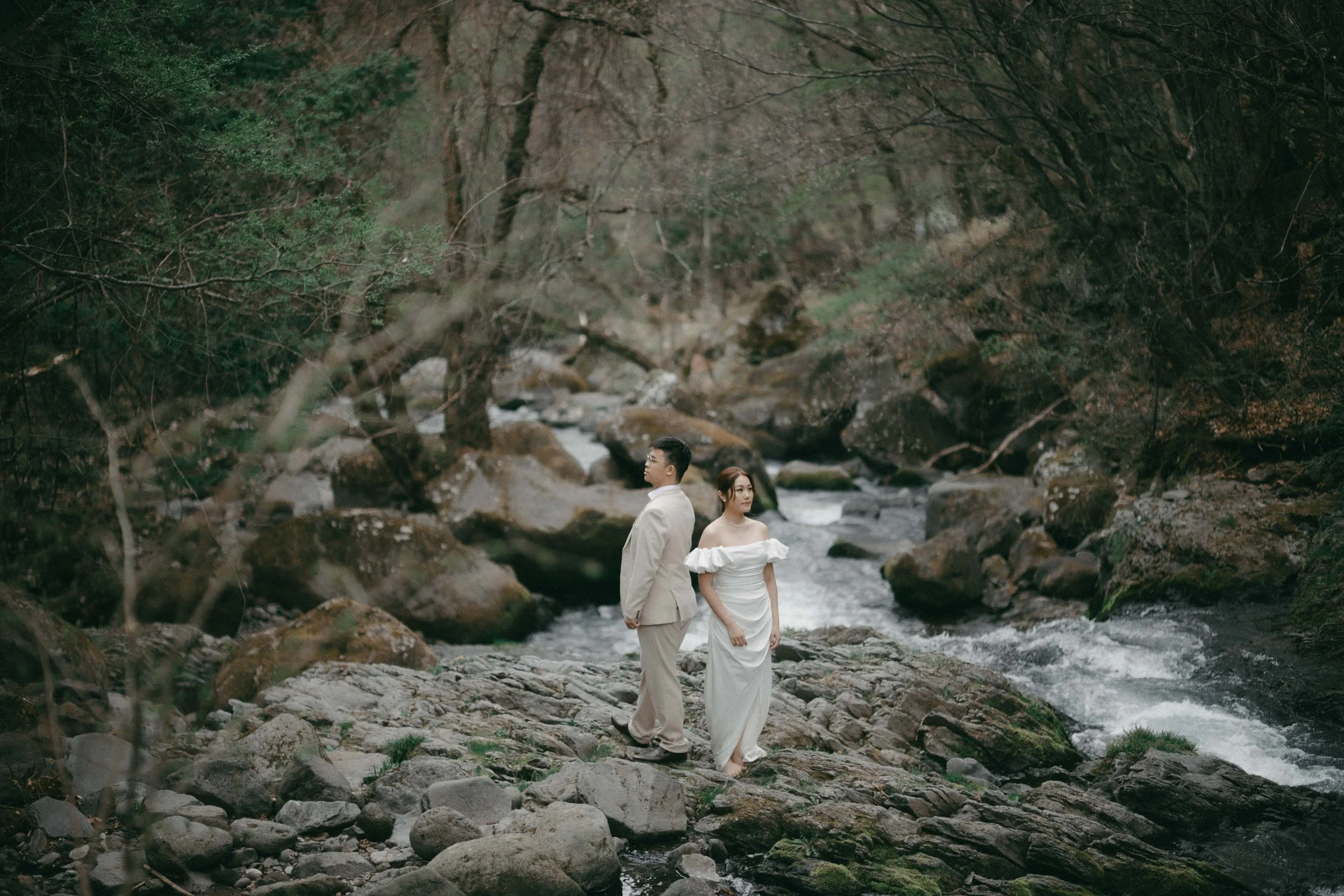 A couple dressed in wedding attire standing on rocks by a flowing river in a forested area with trees and rocks surrounding them.