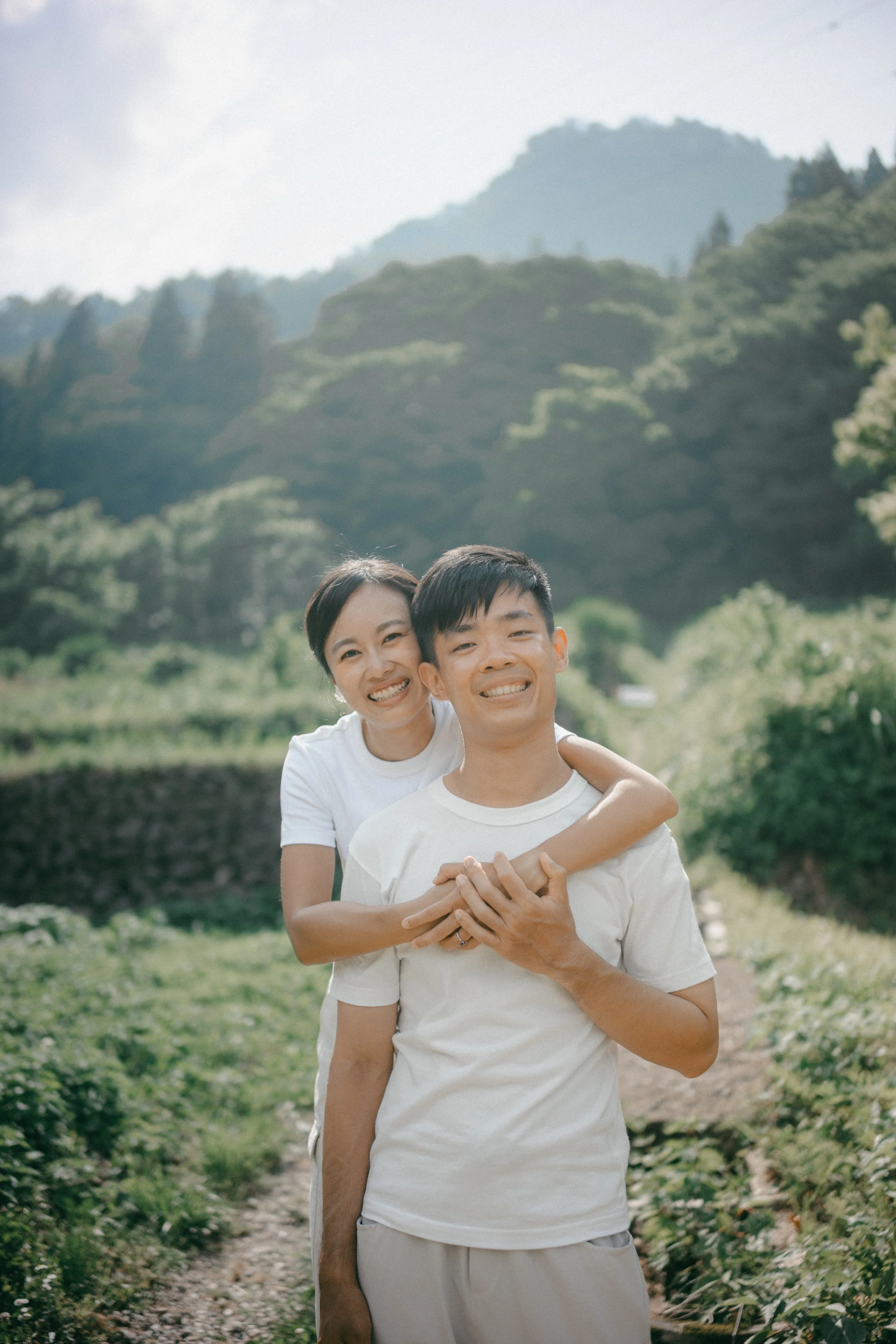 A smiling couple, a woman with arms around a man, standing outdoors in a green, mountainous area on a sunny day.
