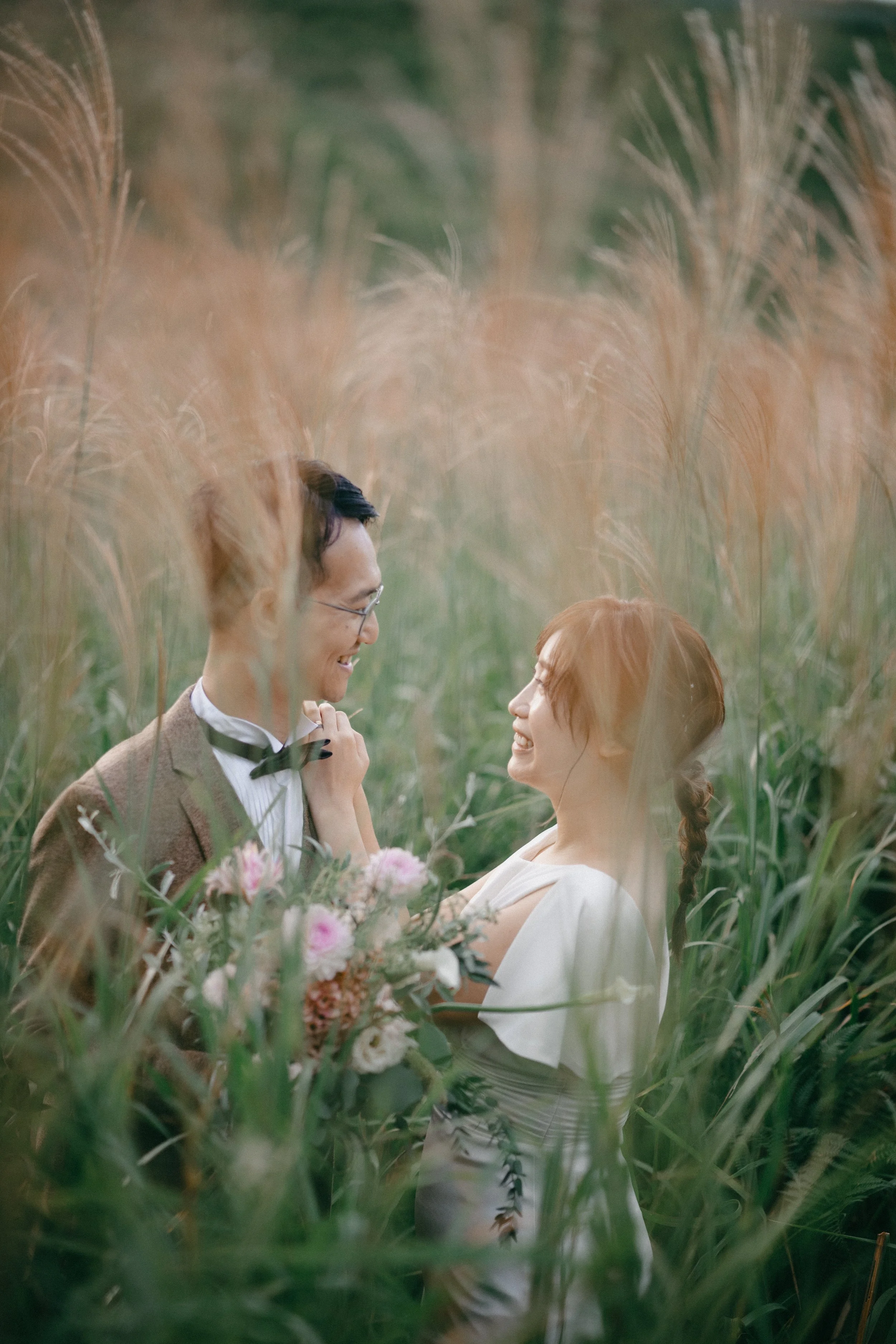 A couple stands close together in a field of tall grass, smiling and gazing at each other. The woman holds a bouquet of pink and white flowers, and the man is dressed in a suit with a bowtie.