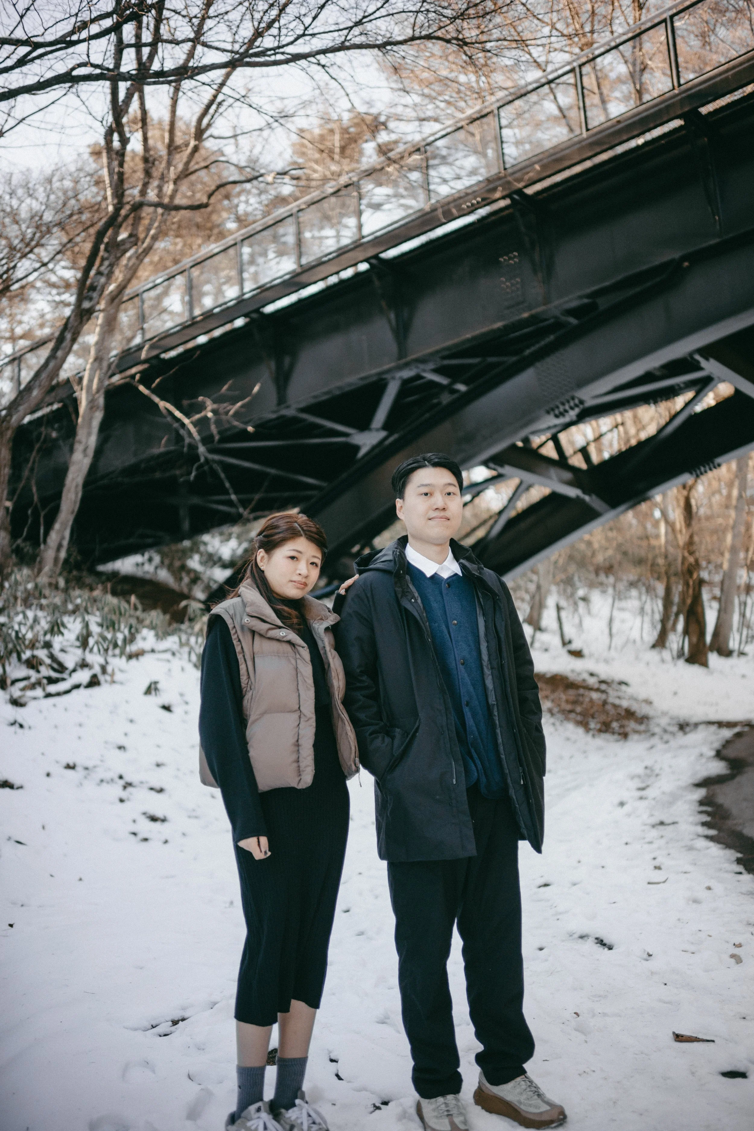 A young woman and man stand in a snowy park under a black bridge, dressed in winter clothing.
