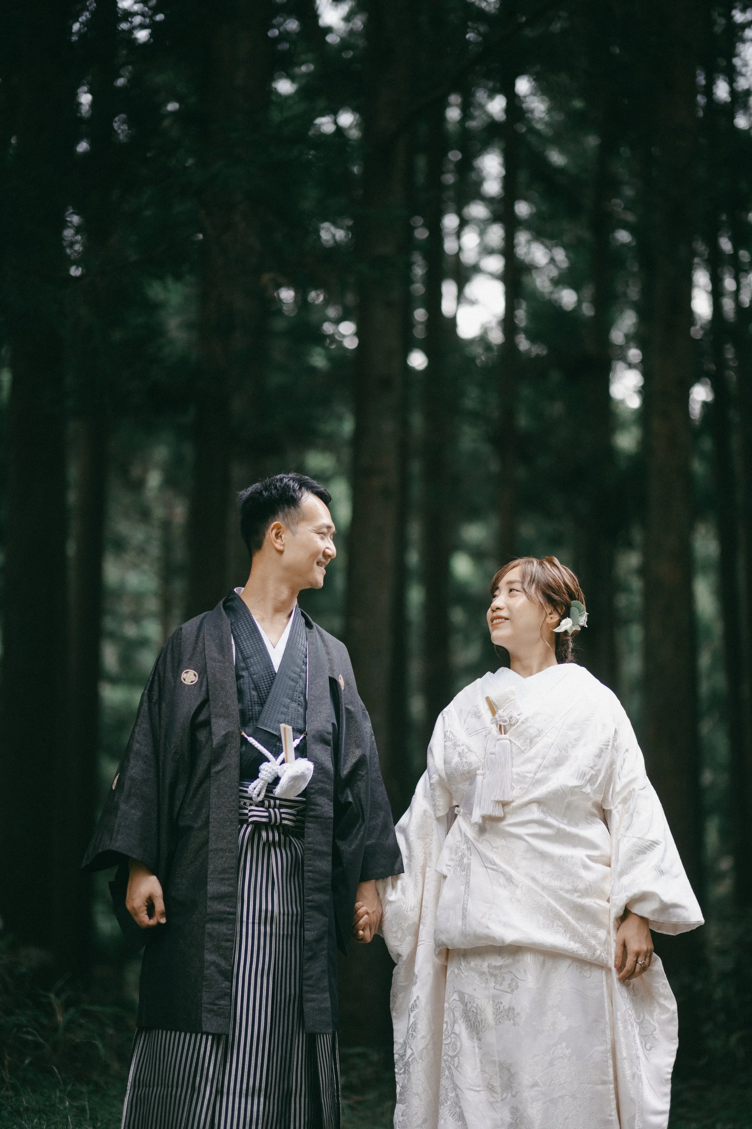 A couple in traditional Japanese wedding attire holding hands and smiling at each other in a forest.