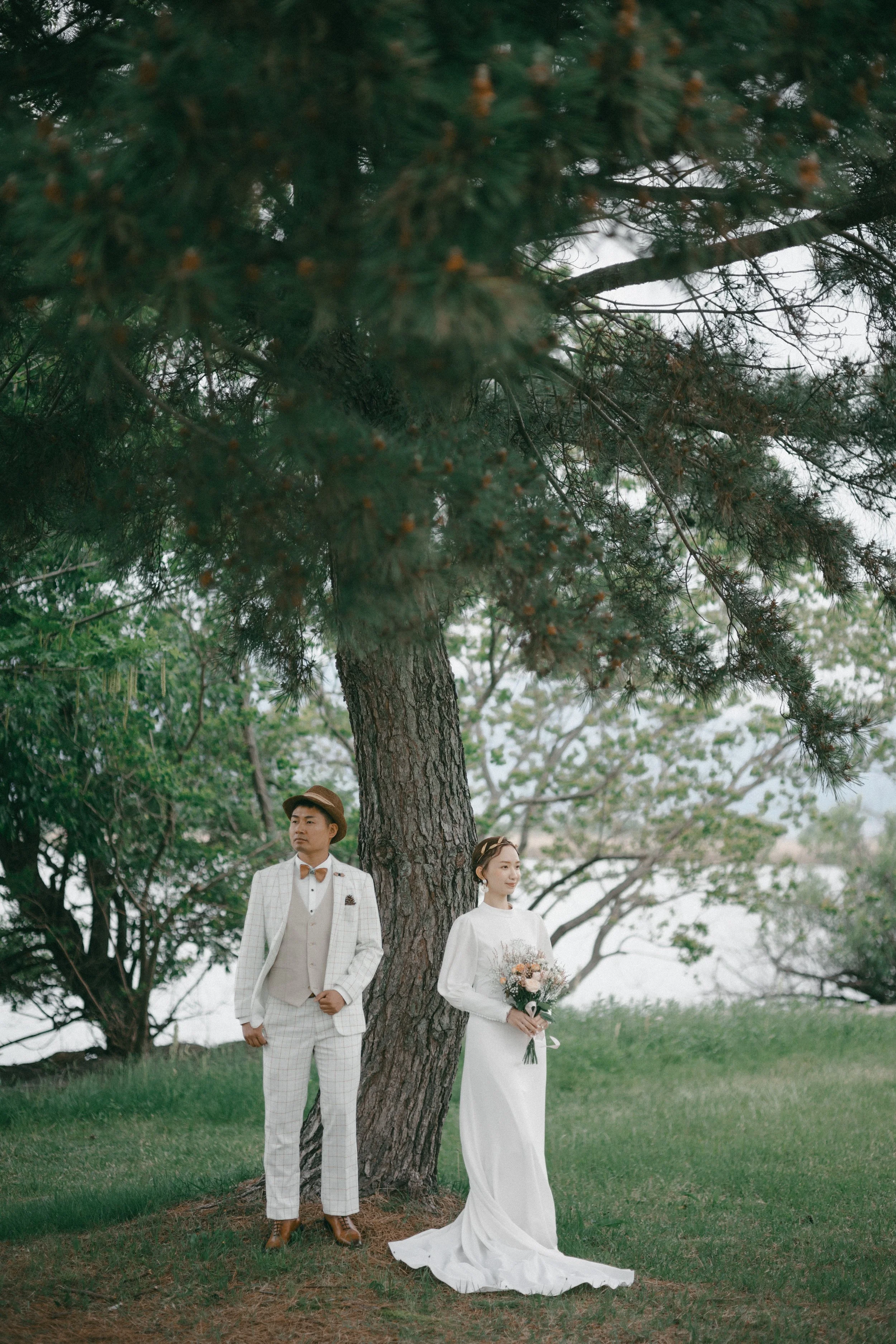 A bride and groom standing outdoors next to a tree, with the bride holding a bouquet of flowers and the groom dressed in a white suit with a brown hat, in a natural, lush setting.