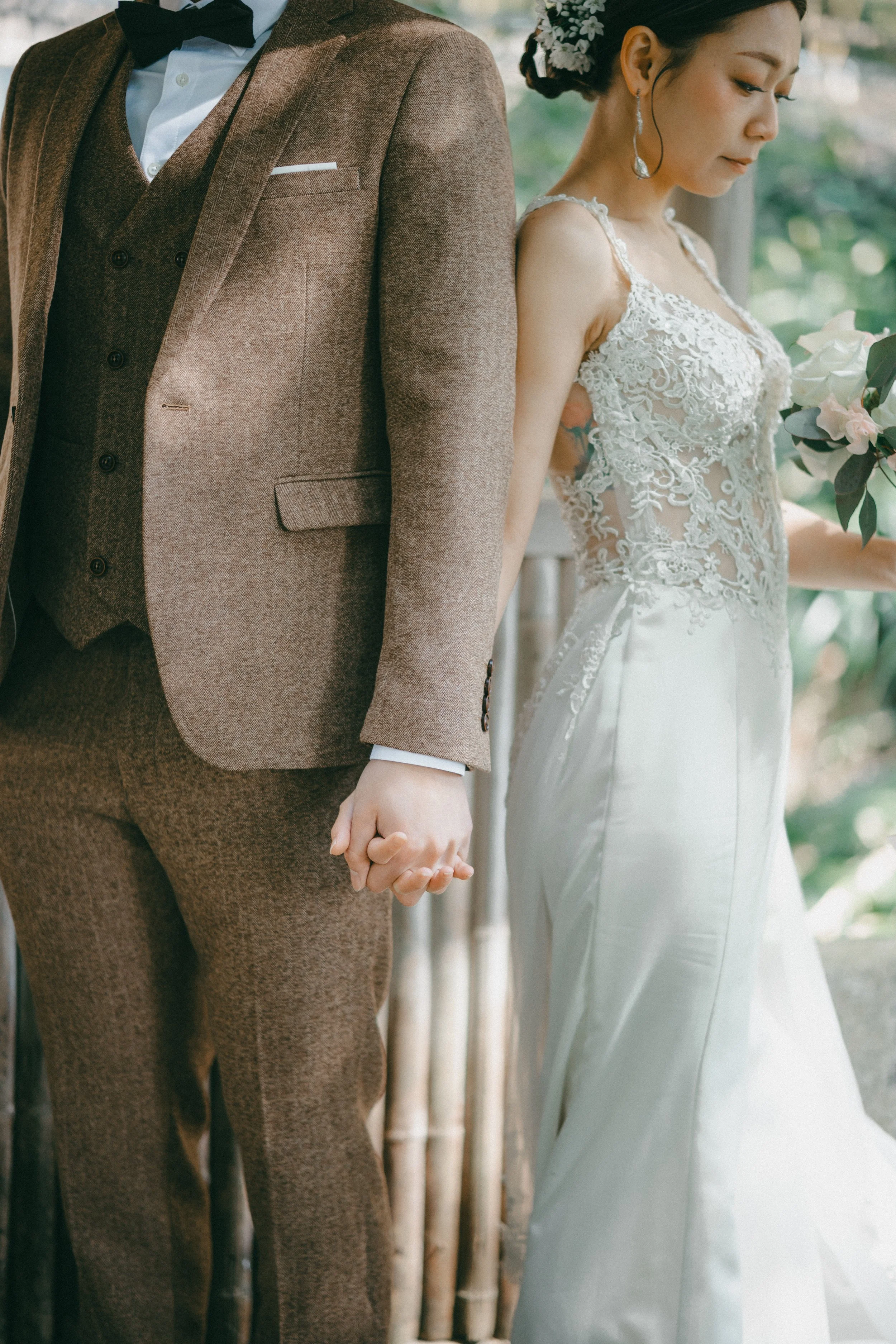 A bride and groom stand side by side during their wedding, holding hands with their backs touching. The bride is wearing a lace wedding gown with intricate floral designs and a floral hair accessory. The groom is dressed in a brown suit with a white 