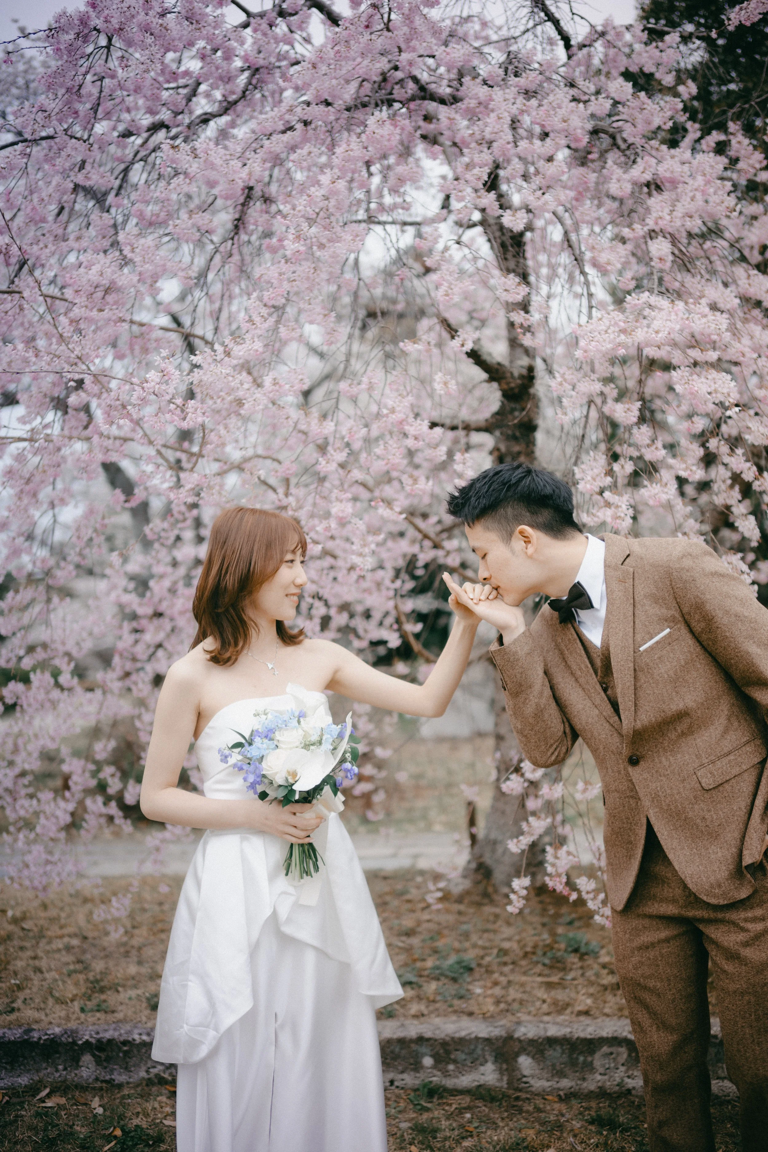 A bride in a white wedding dress holding a bouquet of flowers and a groom in a brown suit with a bowtie, standing under a blooming cherry blossom tree. The groom is kissing the bride's hand.