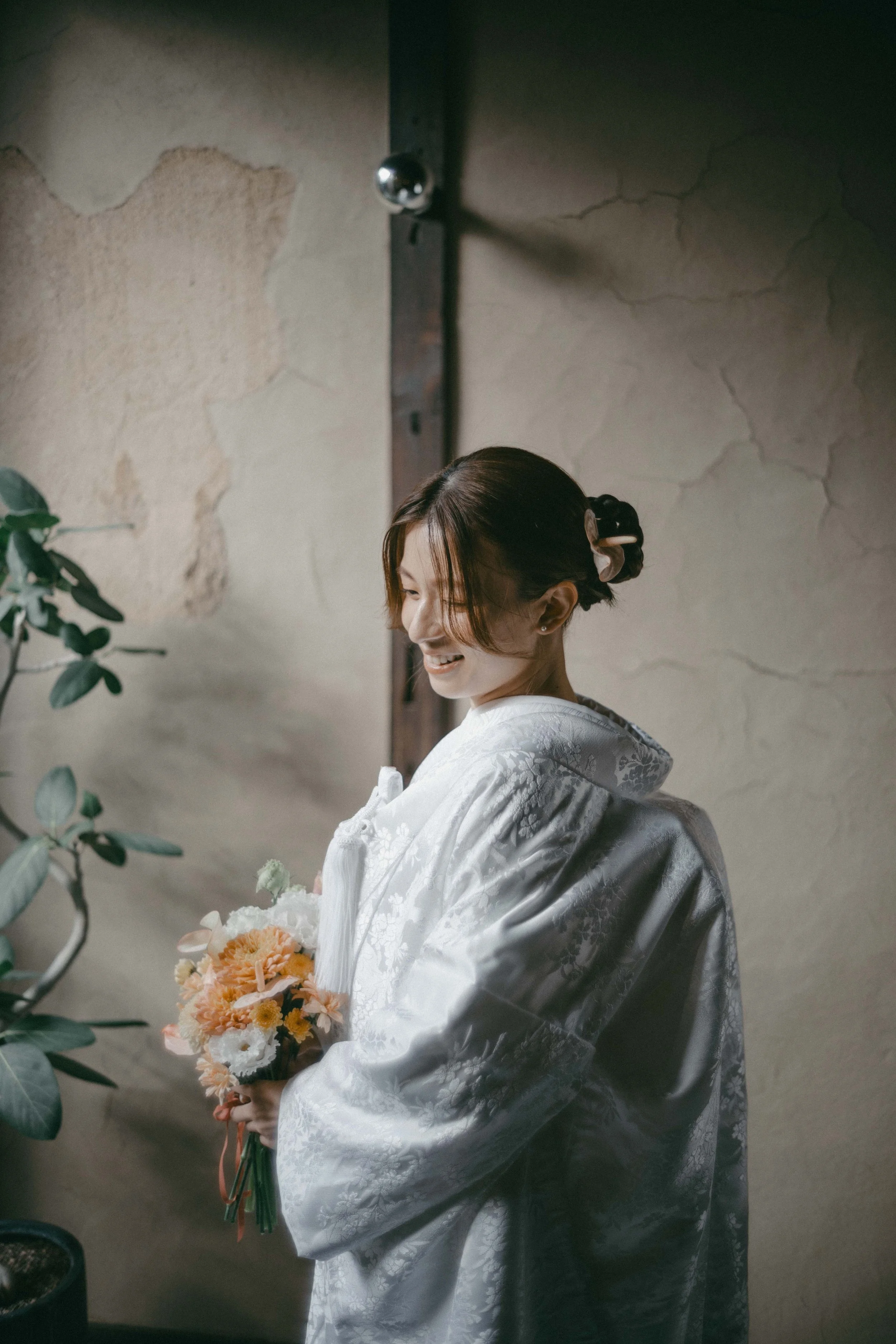 A woman in a white embroidered gown holding a bouquet of peach, white, and orange flowers, standing indoors next to a green plant and a textured wall.