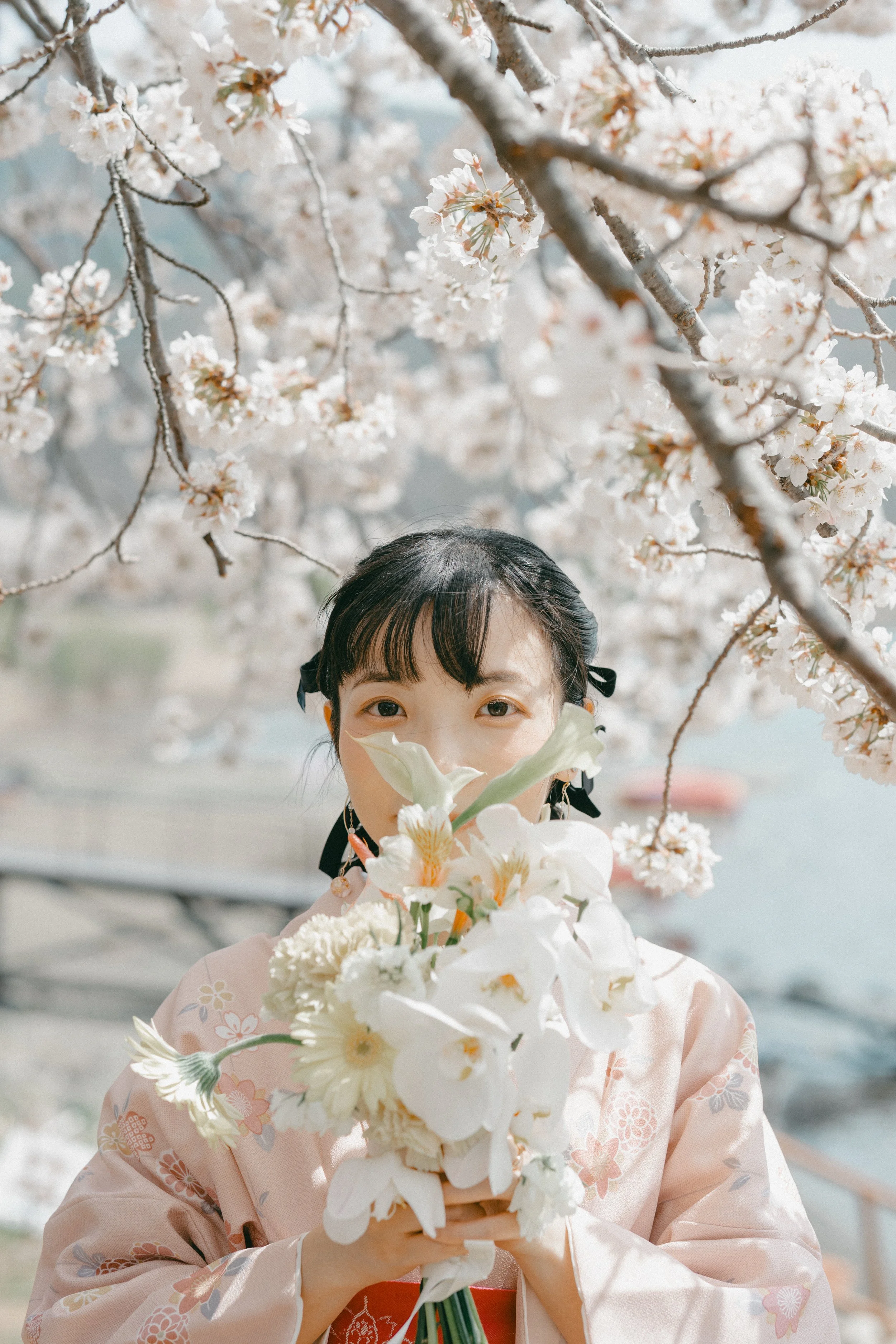 A young woman in a pink floral kimono holding a bouquet of white flowers. She is standing outdoors under cherry blossom trees during spring.